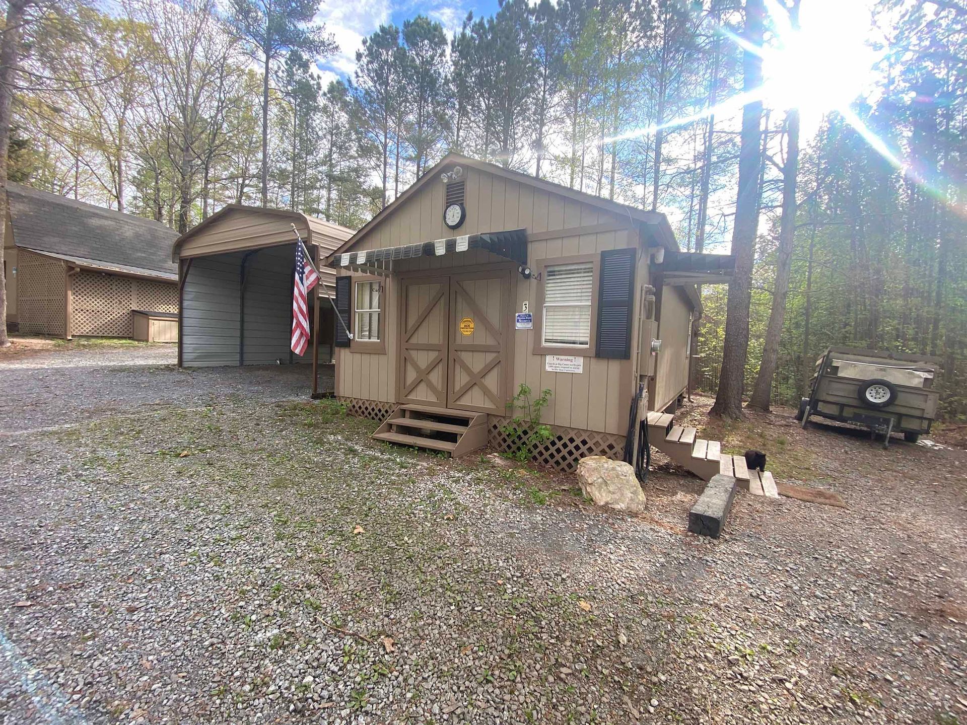 Rustic cabin with small porch and American flag, gravel yard, trees, and ATV in the background.