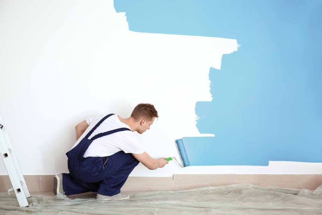 Painter rolling light blue paint on a wall next to a ladder in a room under renovation