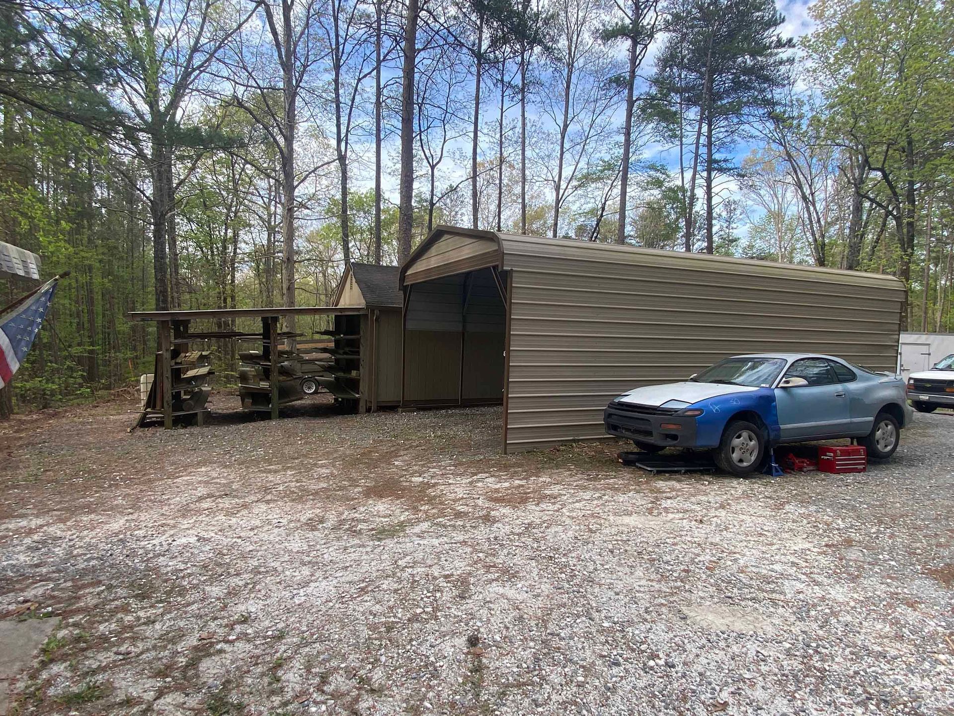 Blue car parked beside a small shed and storage building in a wooded yard
