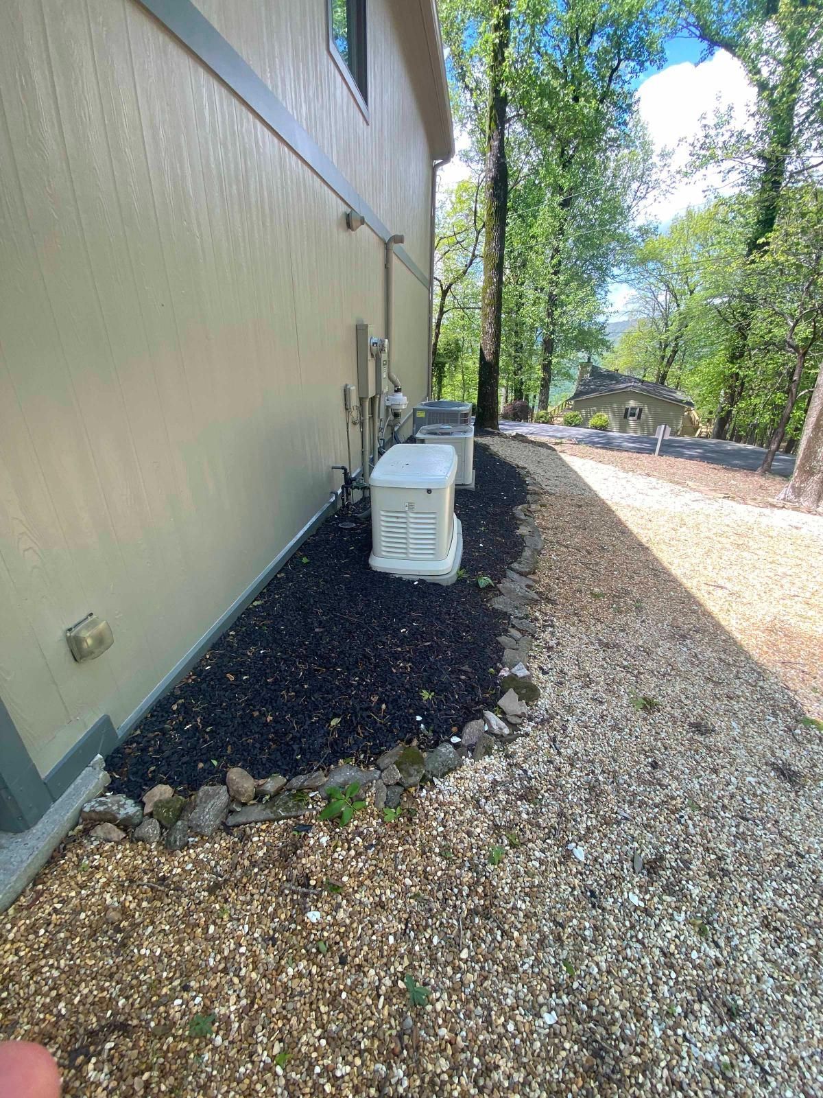 Gravel side yard beside a beige building, with black mulch, utility units, and trees overhead