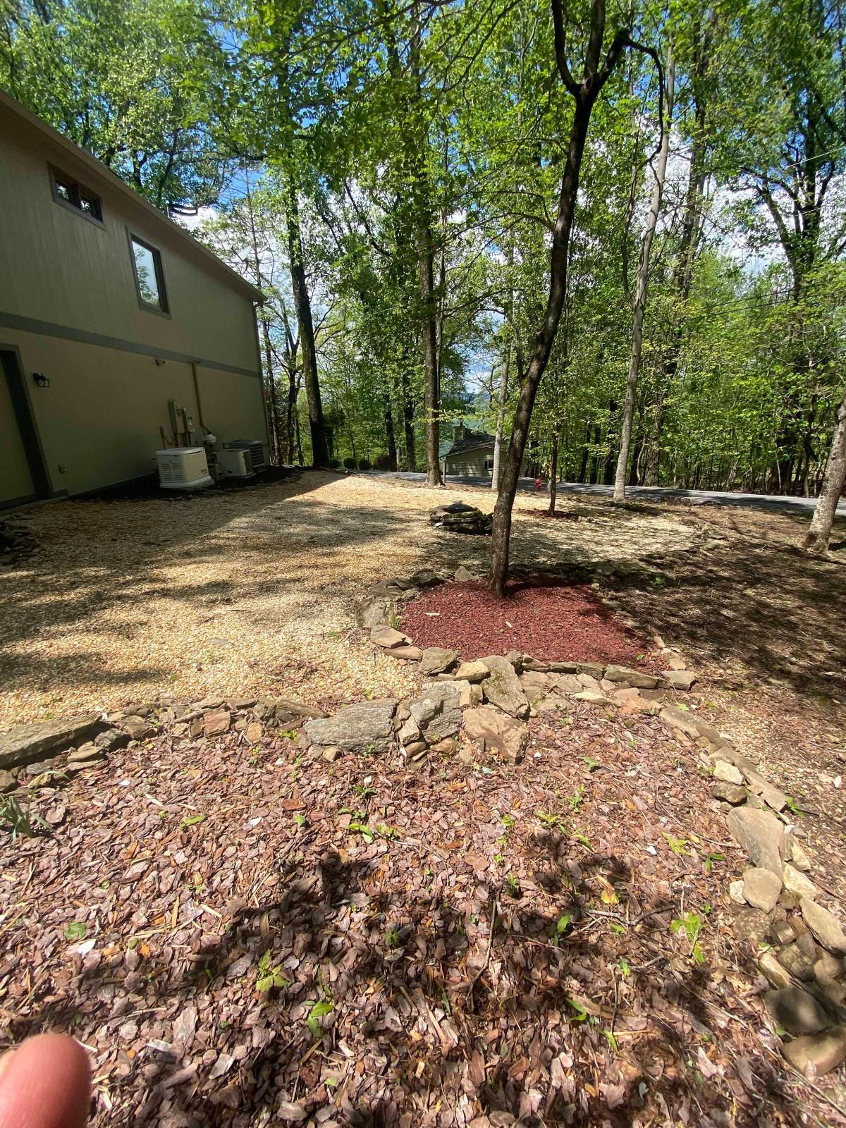 Wooded backyard beside a tan house, with trees, mulch, and a rocky ground cover.