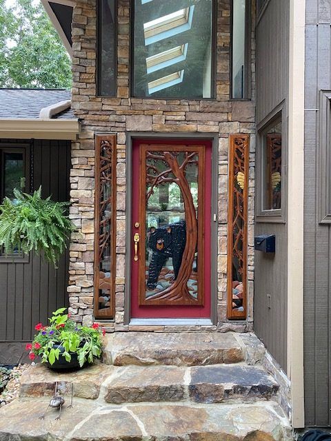 Front entry with stone steps and a red-framed glass door beside potted flowers, reflecting a person outdoors