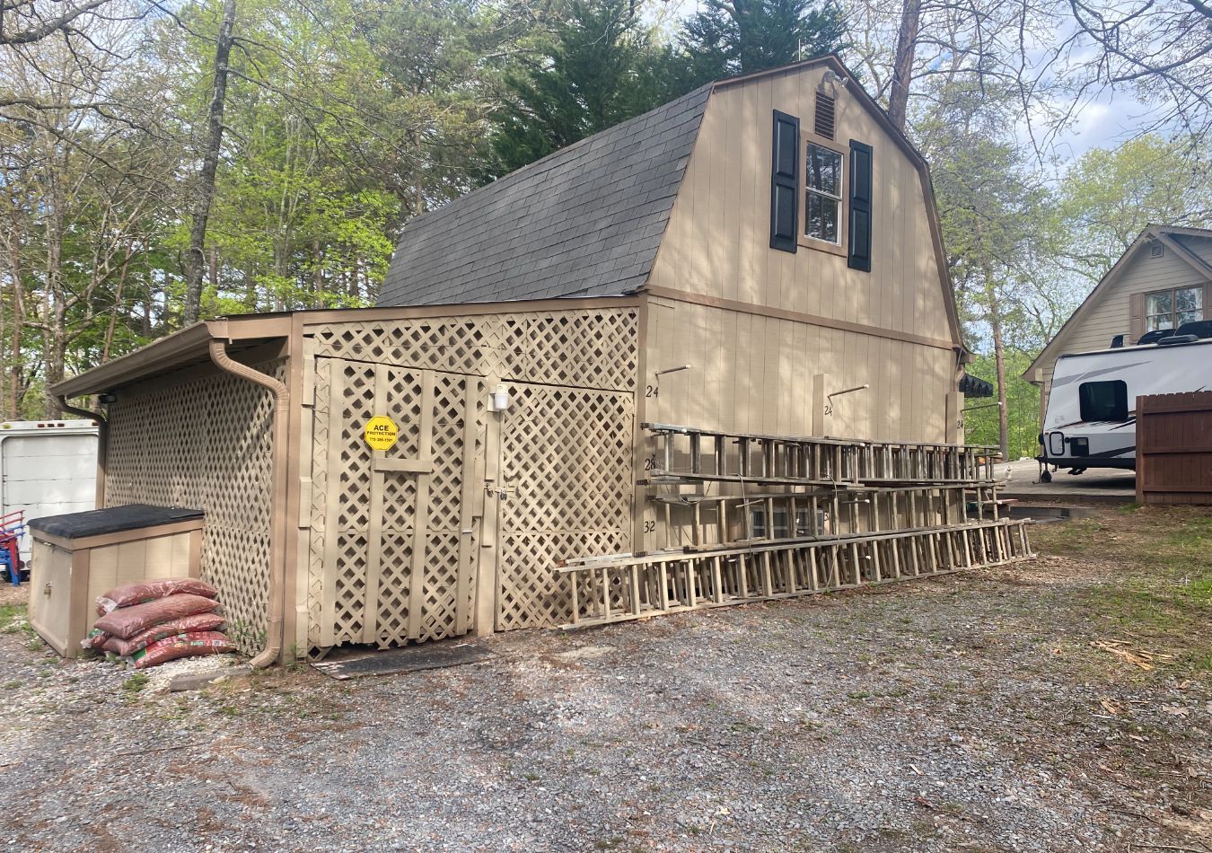 Tan shed with loft windows and lattice skirting beside a gravel driveway and trees