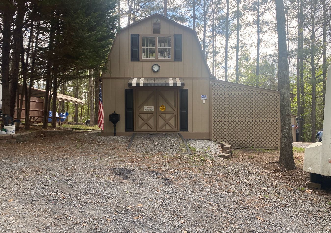 Small tan barn-style cabin with double doors and windows, set among trees on a gravel driveway.