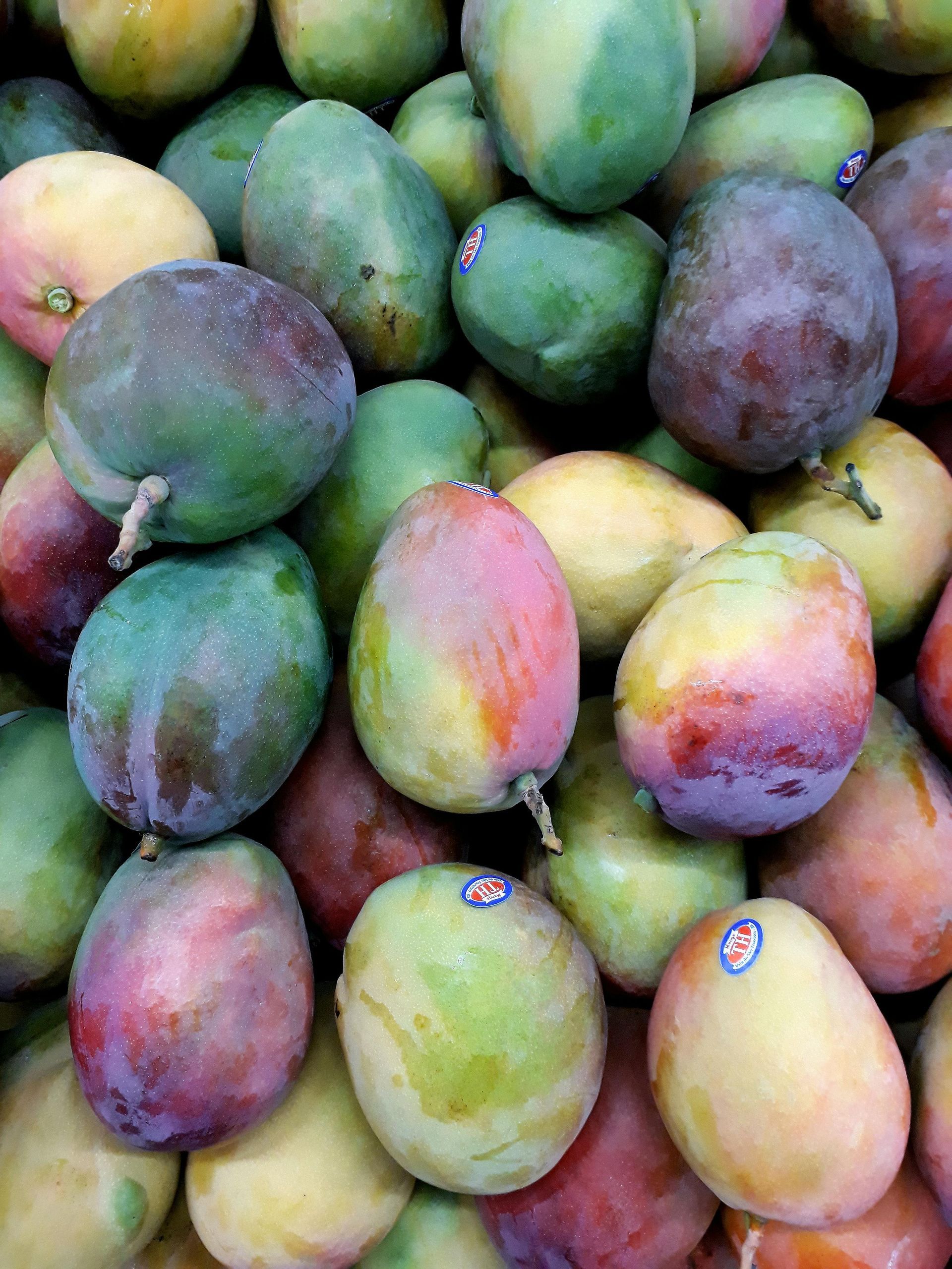 Pile of colorful mangoes: green, yellow, and red skins, some with stickers, close-up shot.