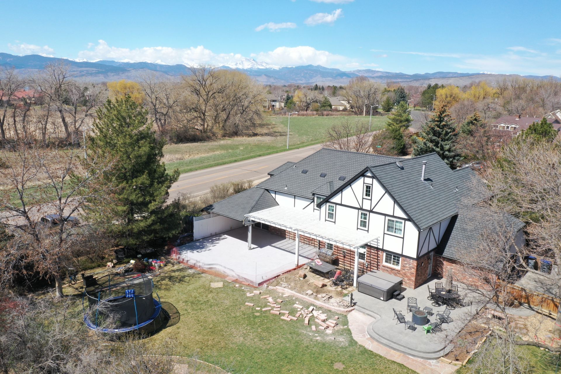 Aerial view of Tudor-style house with black roof, white trim, and red brick. Backyard has a trampoline, patio, and fire pit. Mountains in the distance.