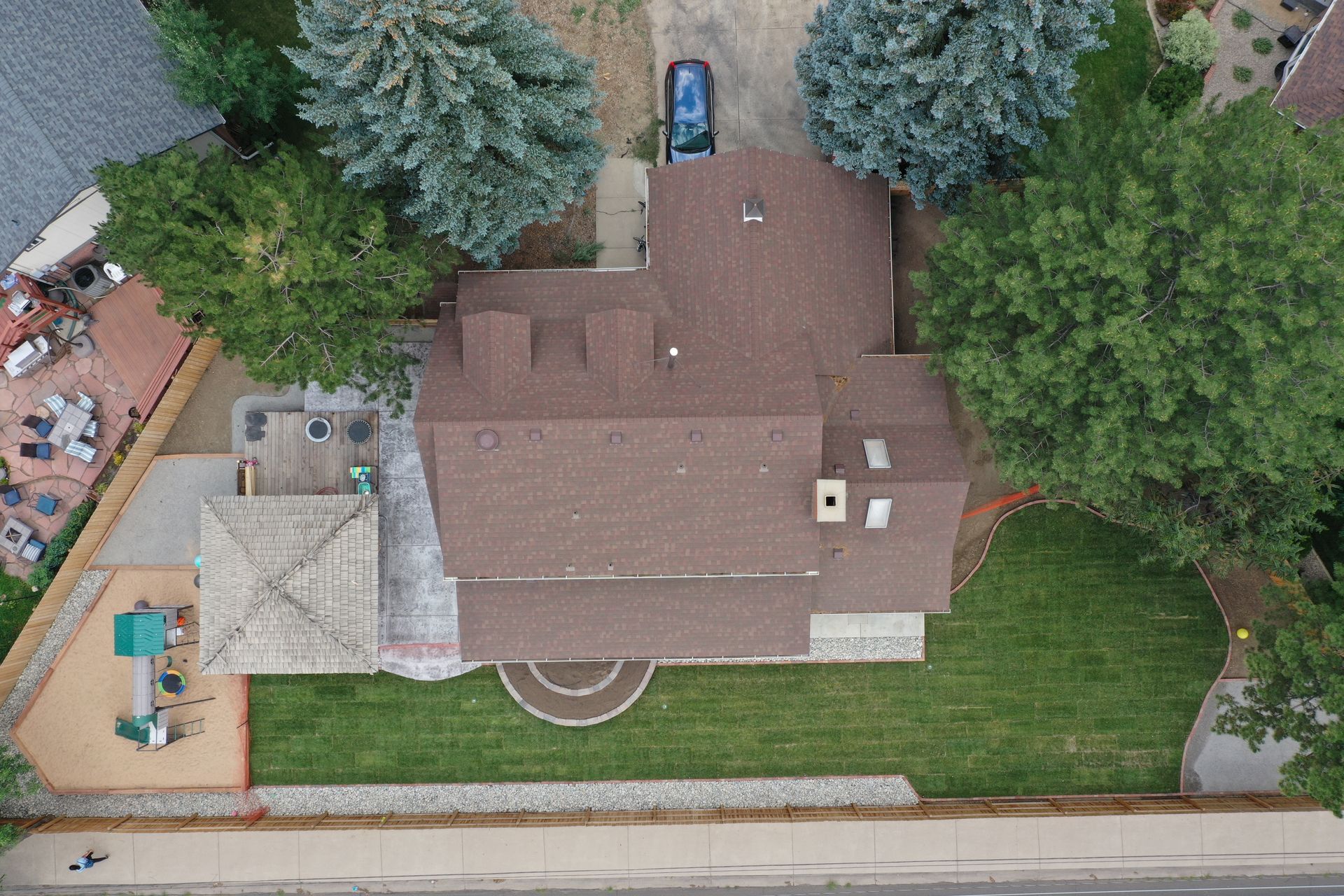 Aerial view of house with brown roof, green lawn, trees, and a car in the driveway.
