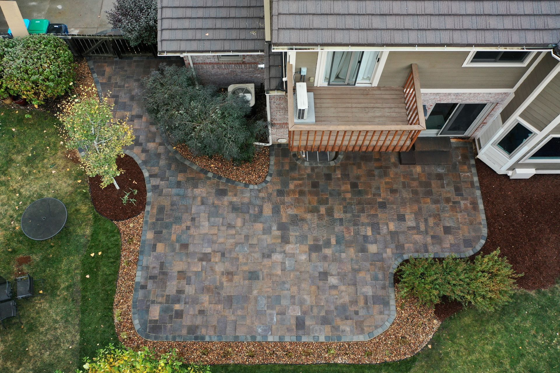 Overhead view of a patio with a brick pattern, bordered by landscaping and a house with a deck.