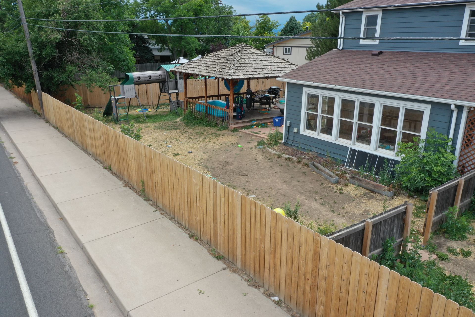 A wooden fence lines a sidewalk next to a house with a gazebo and yard.