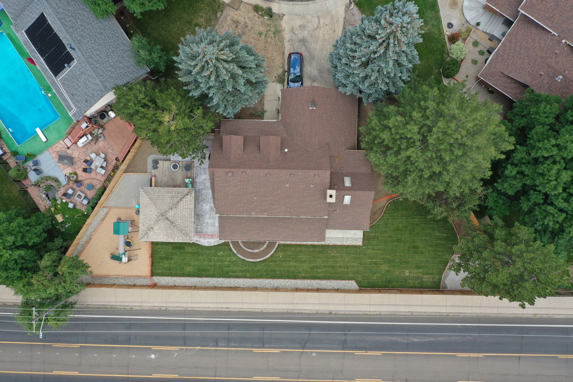 Aerial view of a house with a brown roof, green lawn, pool, and a car in the driveway, near a road.