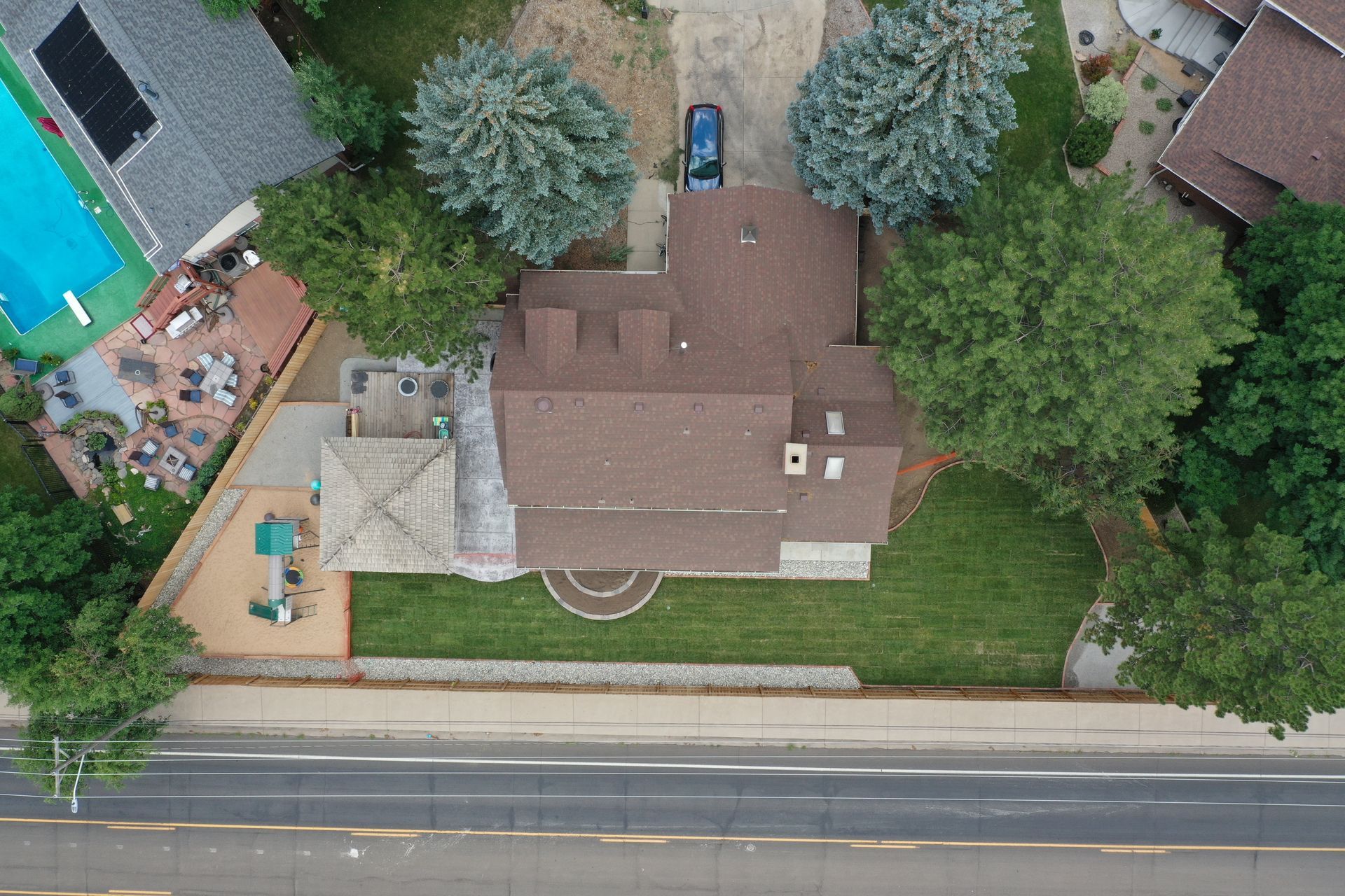 Aerial view of a house with a brown roof, green lawn, pool, and a road.