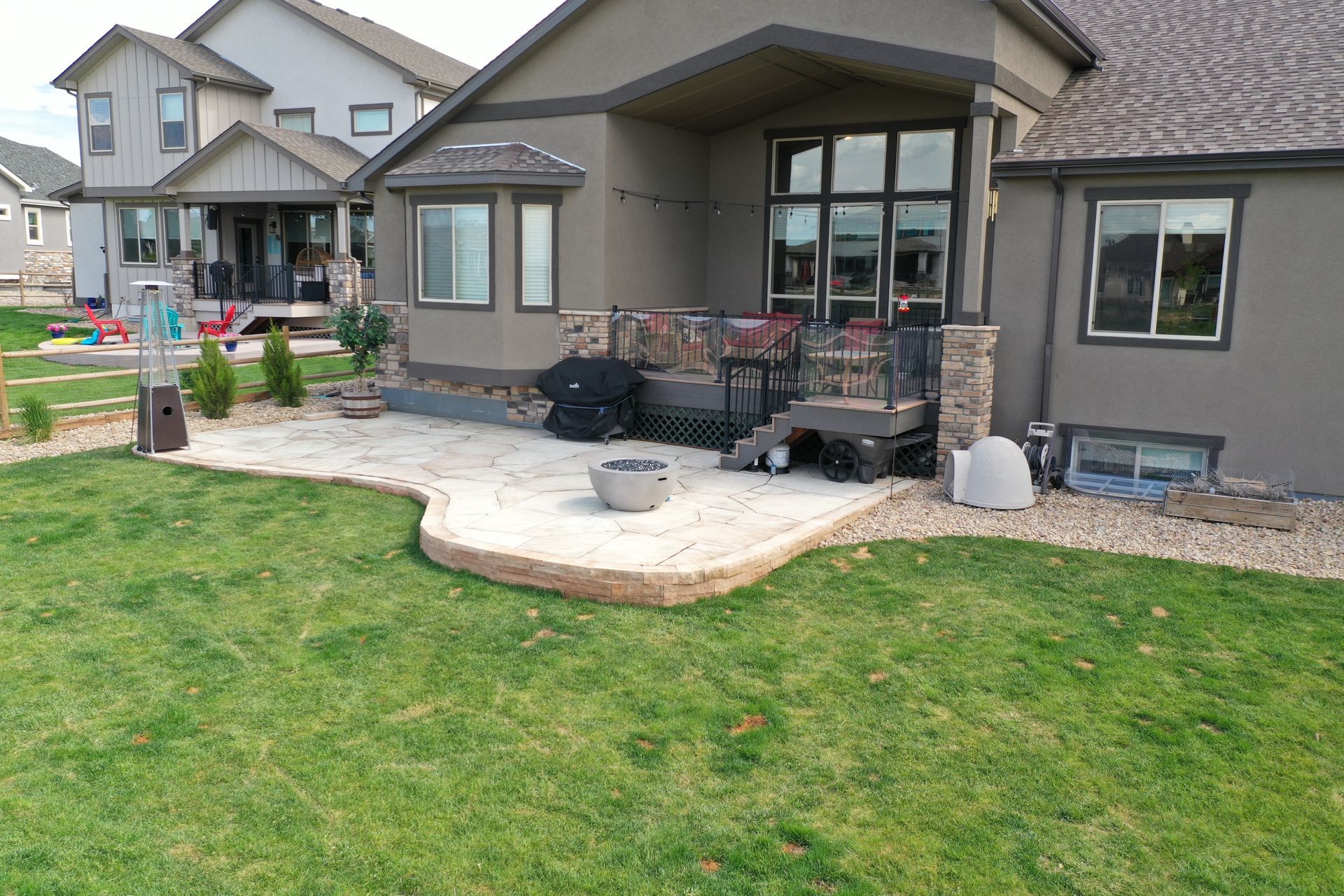 A backyard patio with a grill, fire pit, and seating. Green grass surrounds the patio of a beige house.
