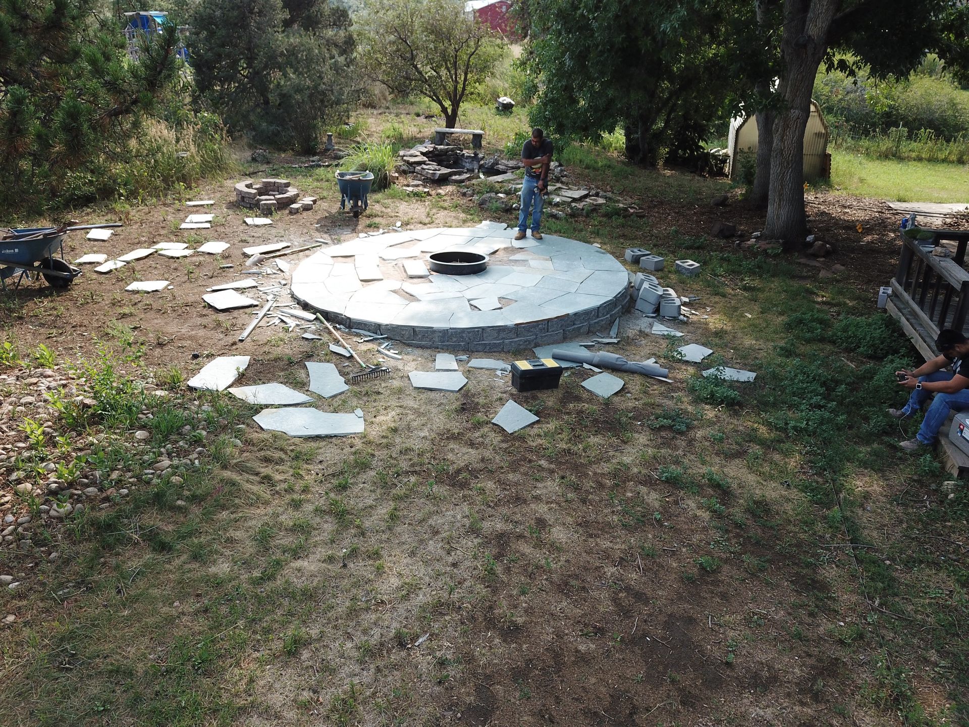 People tiling a circular stone patio outdoors, surrounded by grass, trees and debris.
