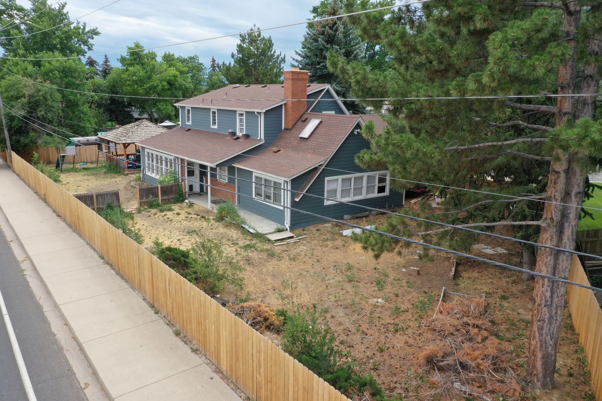 A two-story blue house with a brown roof and a wooden fence along a road.