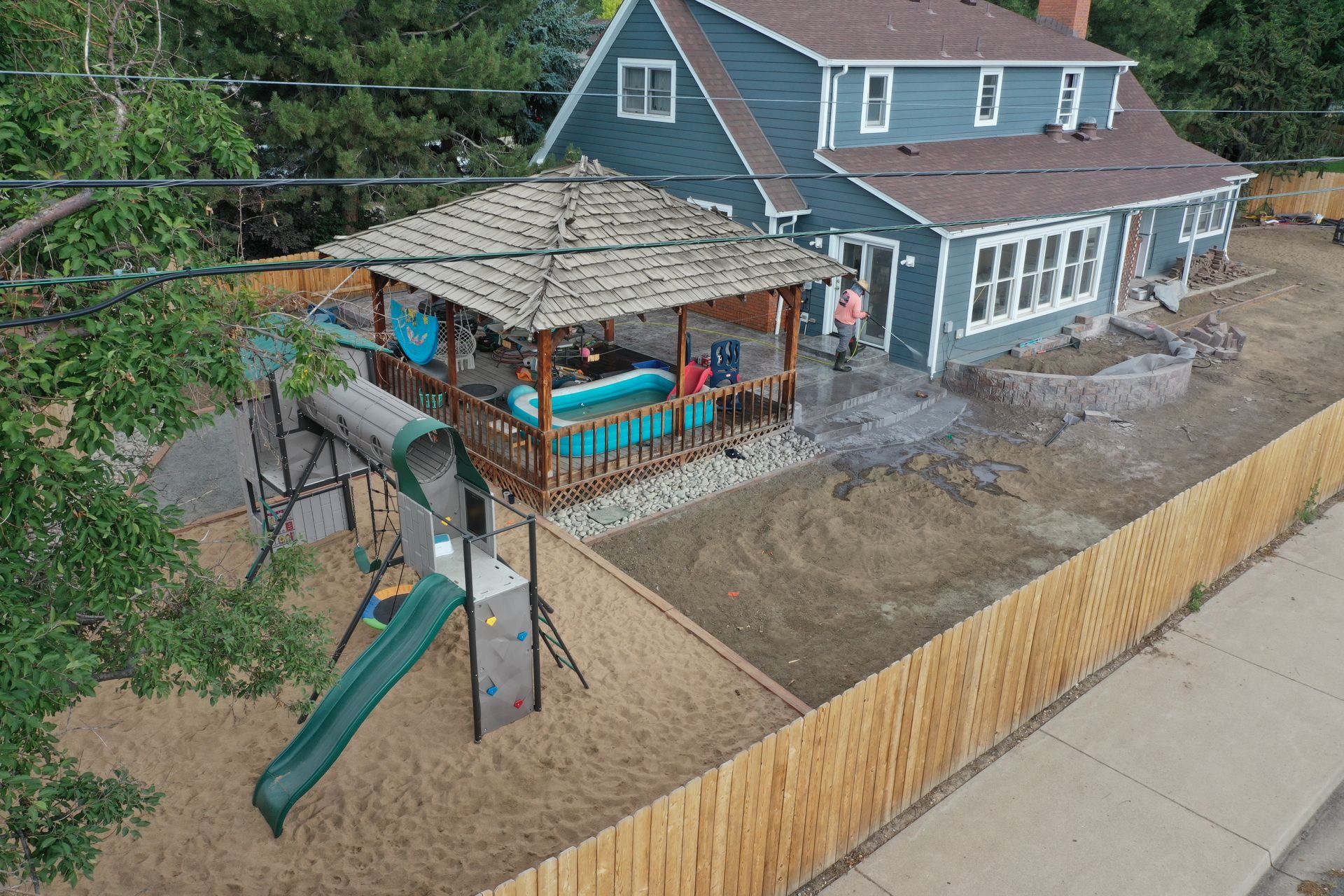 Backyard with a blue house, wooden gazebo, and playground.  Wooden fence surrounds sand play area.