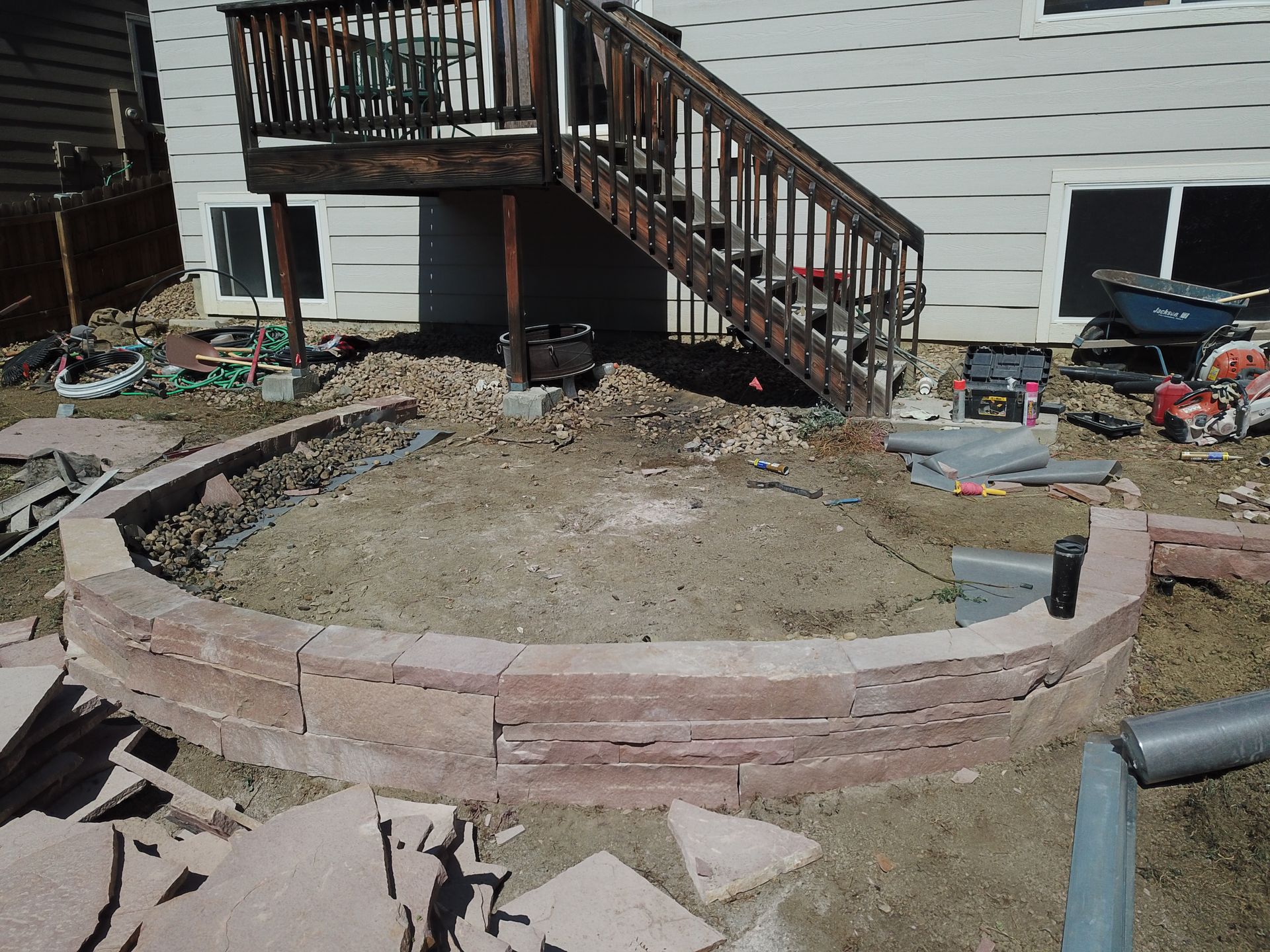 A partially constructed circular patio with red brick walls, beneath a wooden deck, near a house.