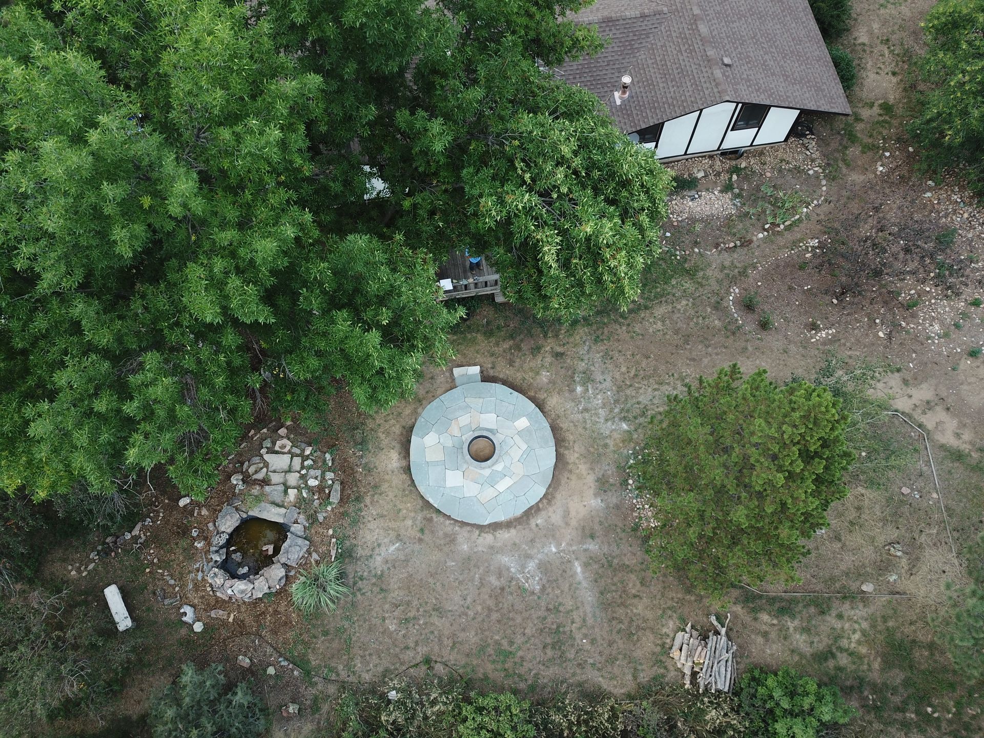 Aerial view: Stone circle, house with brown roof, trees, small pond, and firewood.