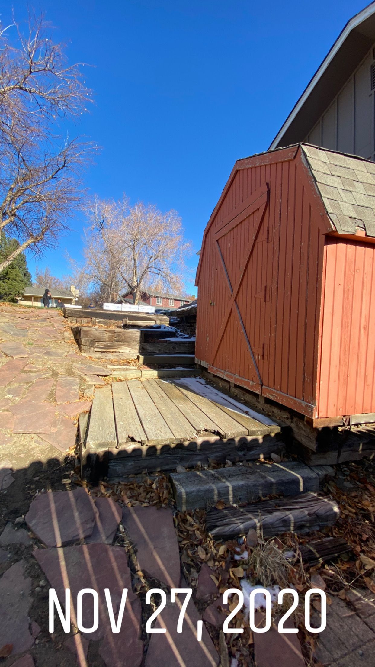 Red shed on brick base, wooden walkway, blue sky, and tree on a sunny day.