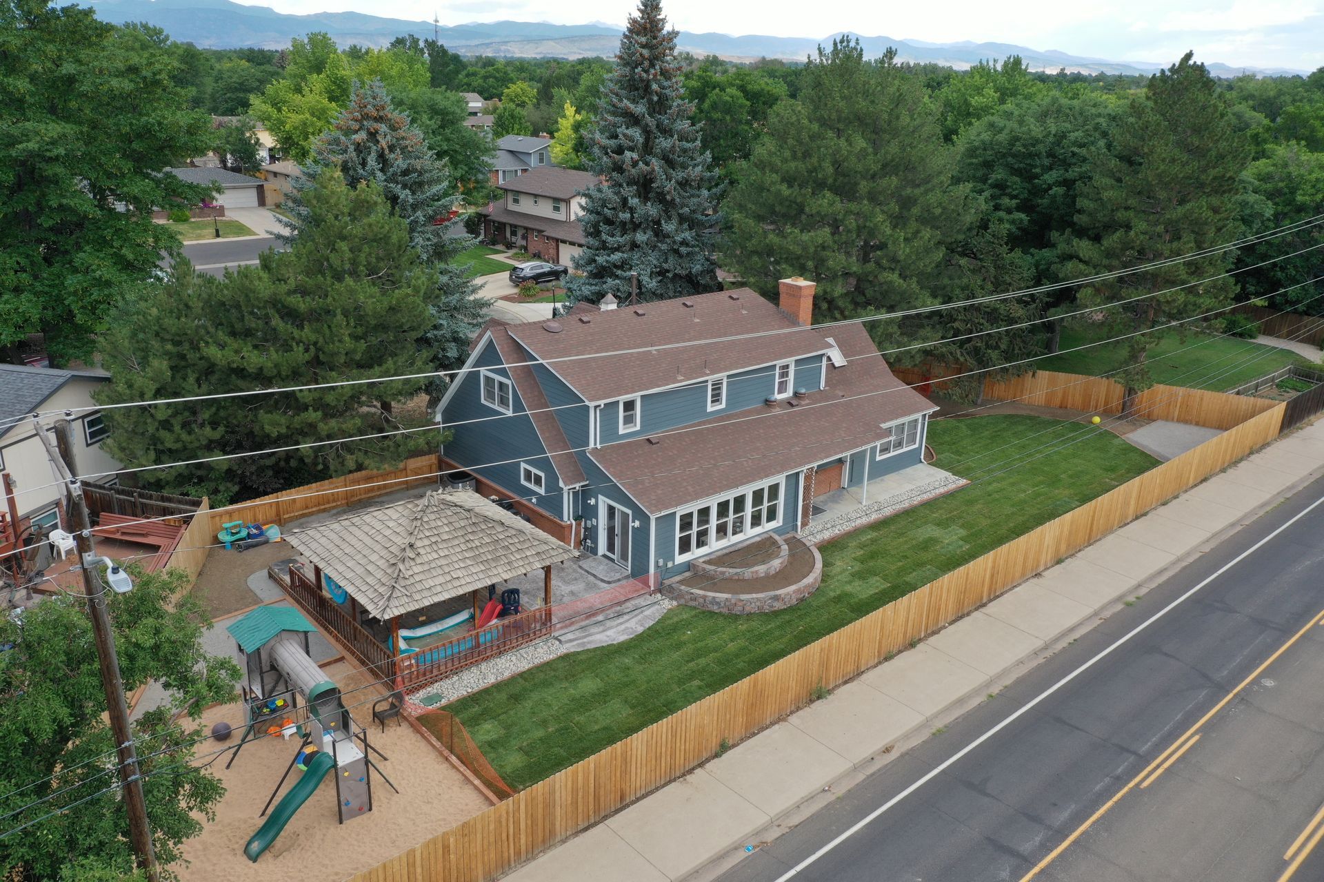 An aerial view of a two-story blue house with a brown roof and a wooden fence. A playset is in the yard.