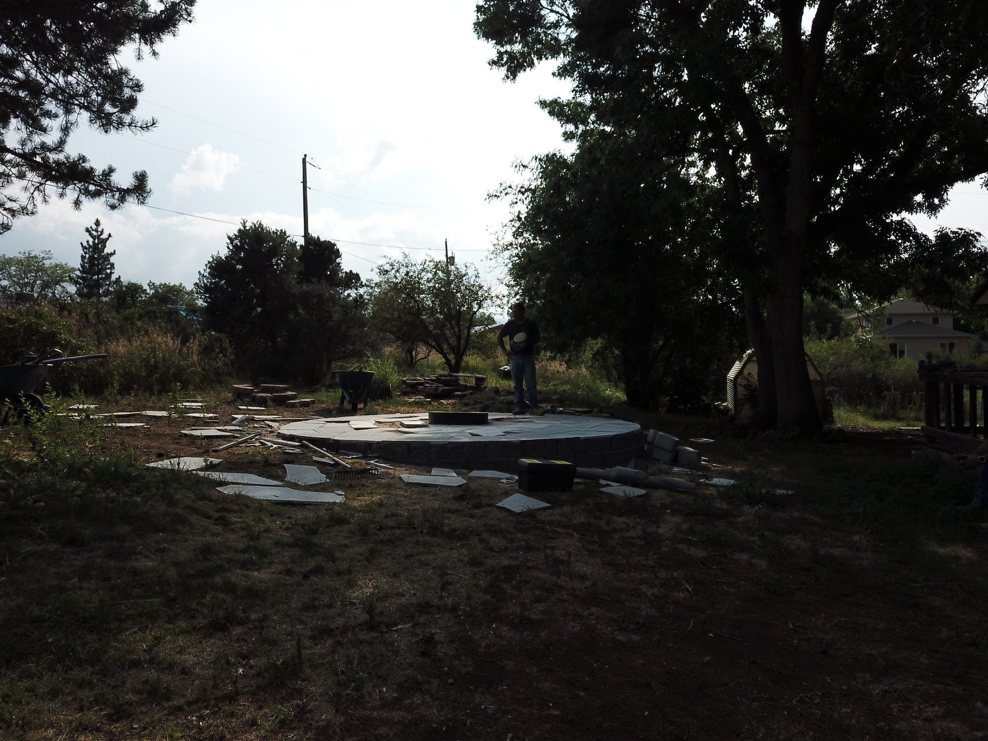Person standing on a round concrete structure outdoors, surrounded by debris, trees, and tall grass on a sunny day.