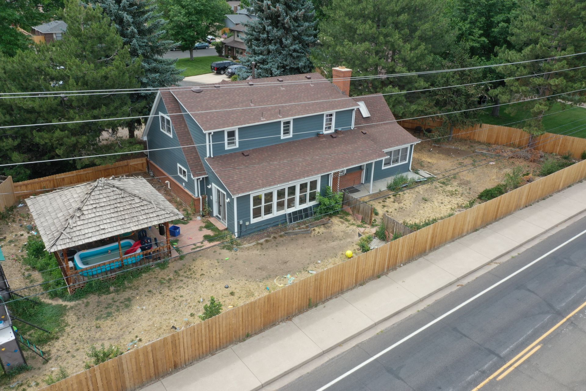 Two-story blue house with a brown roof and a gazebo, surrounded by a wooden fence next to a road.