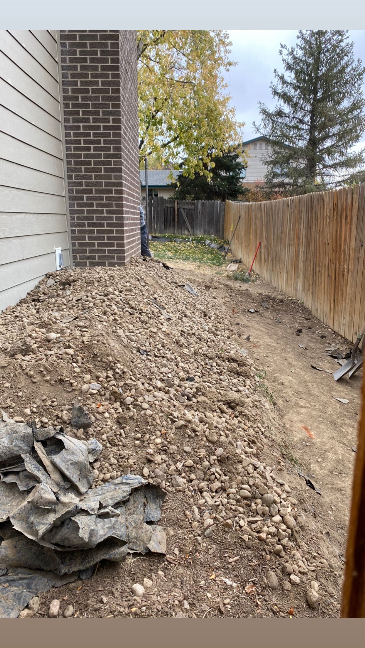 Pile of dirt next to a brick chimney, wooden fence, and siding. Overcast sky.