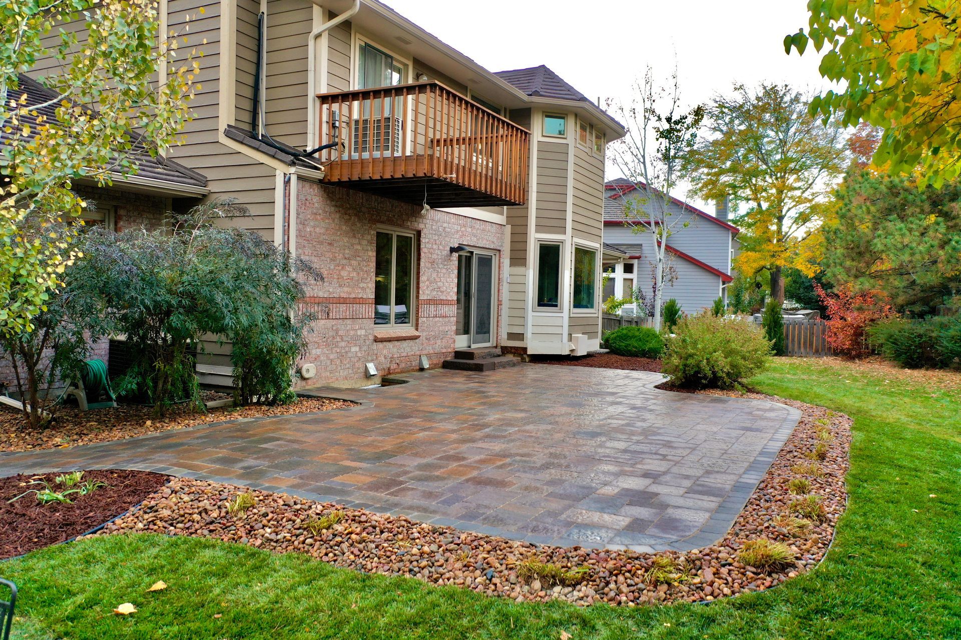 Backyard patio with brick pavers, surrounded by landscaping, beside a multi-story house with a wooden balcony.