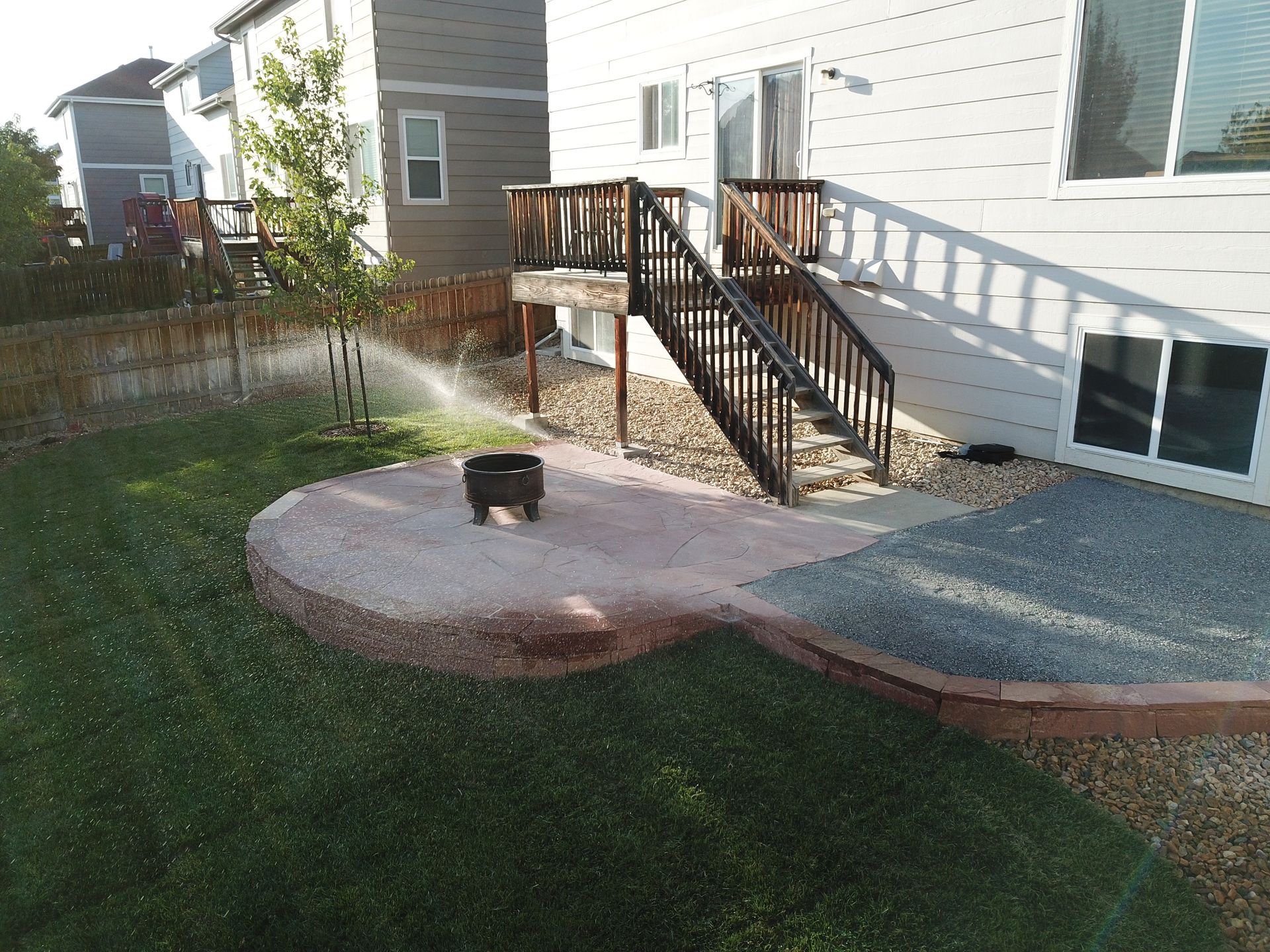 Backyard with a grassy lawn, stone patio, stairs leading up to a deck, and an active sprinkler.