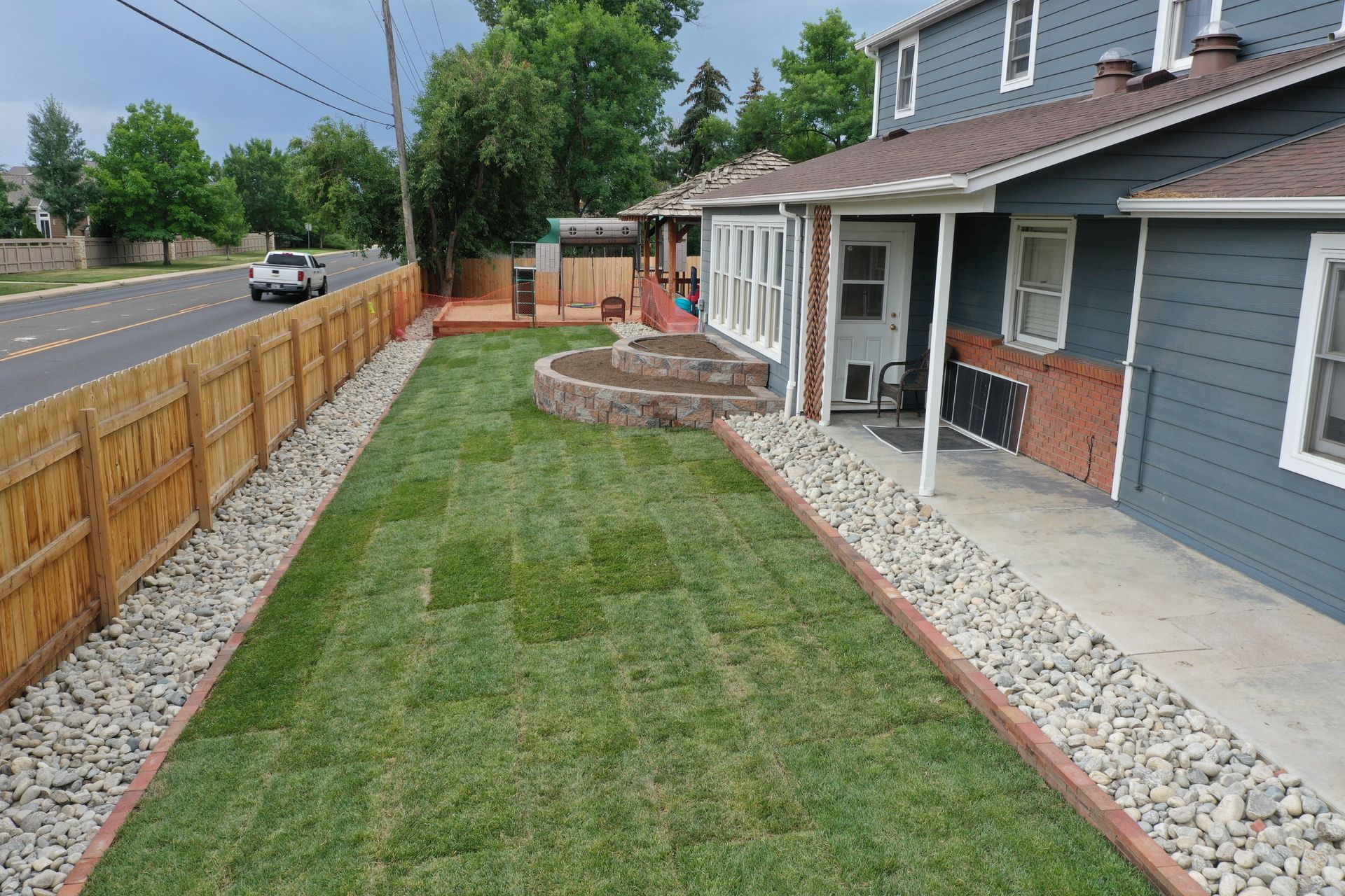 Lawn with fresh sod, bordered by gravel and a wooden fence, alongside a house with a porch.