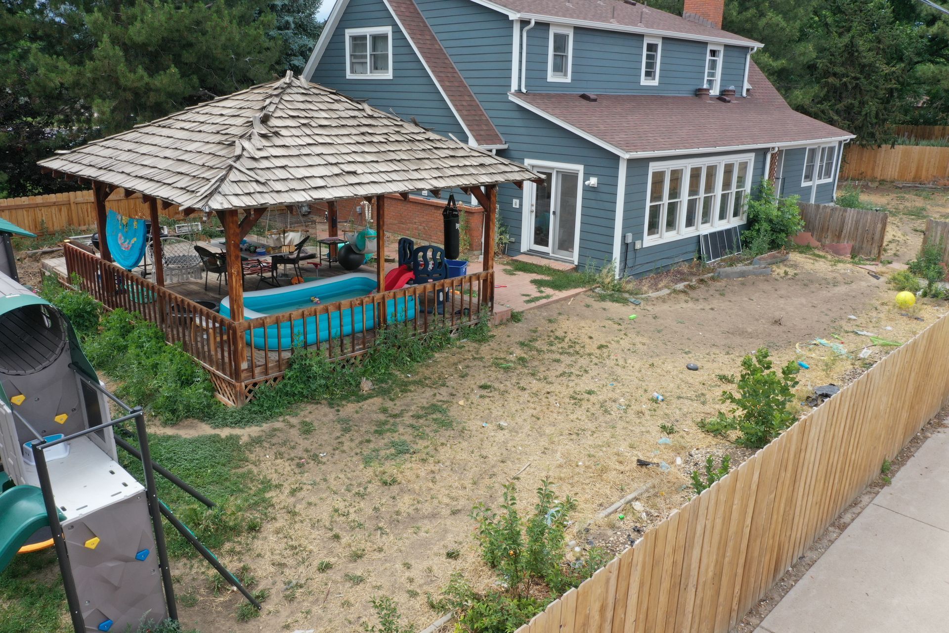 Backyard scene with a house, gazebo, pool, and playground. Wooden fence, dry grass, and blue siding.