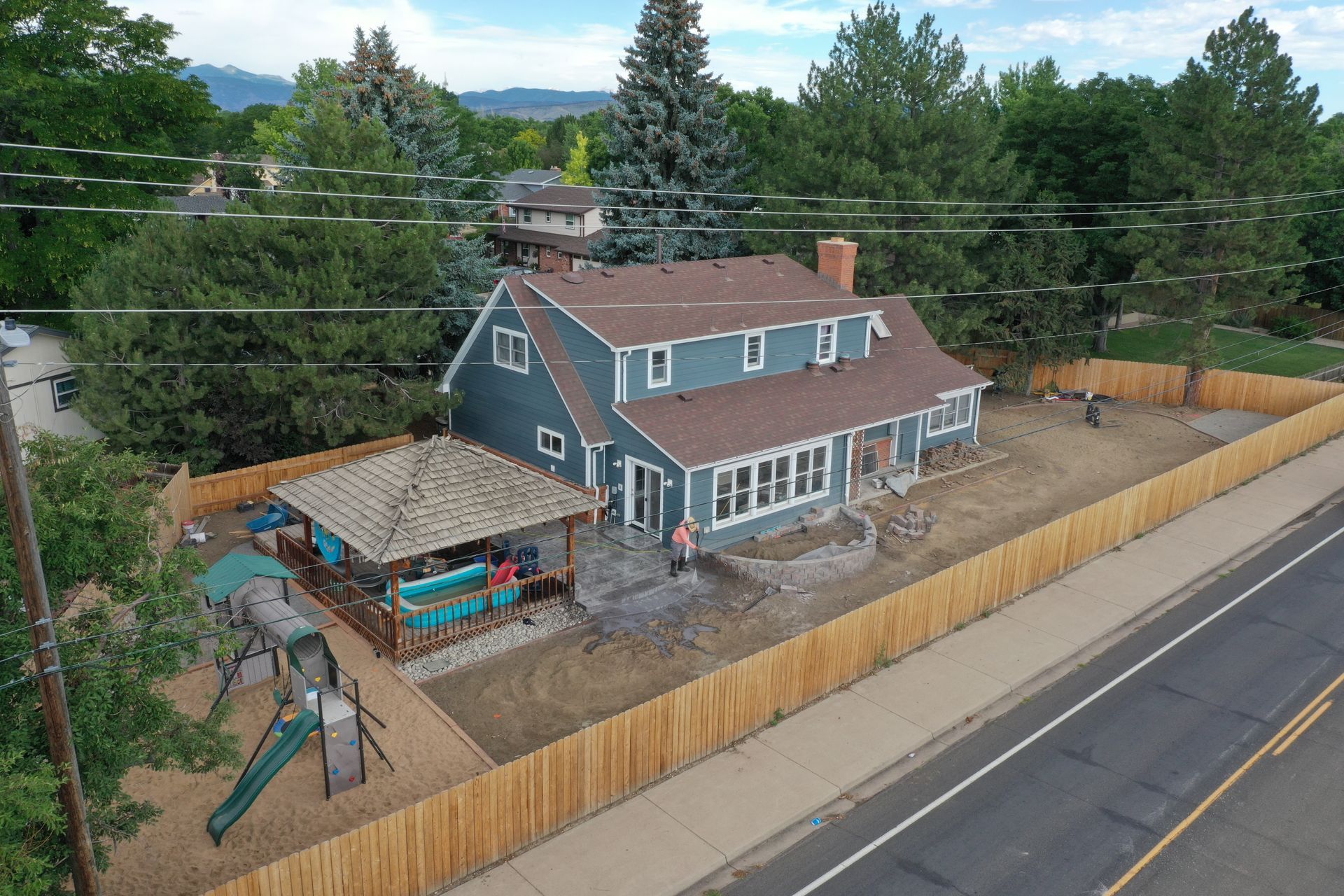 Blue house with brown roof, wooden fence, gazebo, and playground. Trees and mountains in the background.