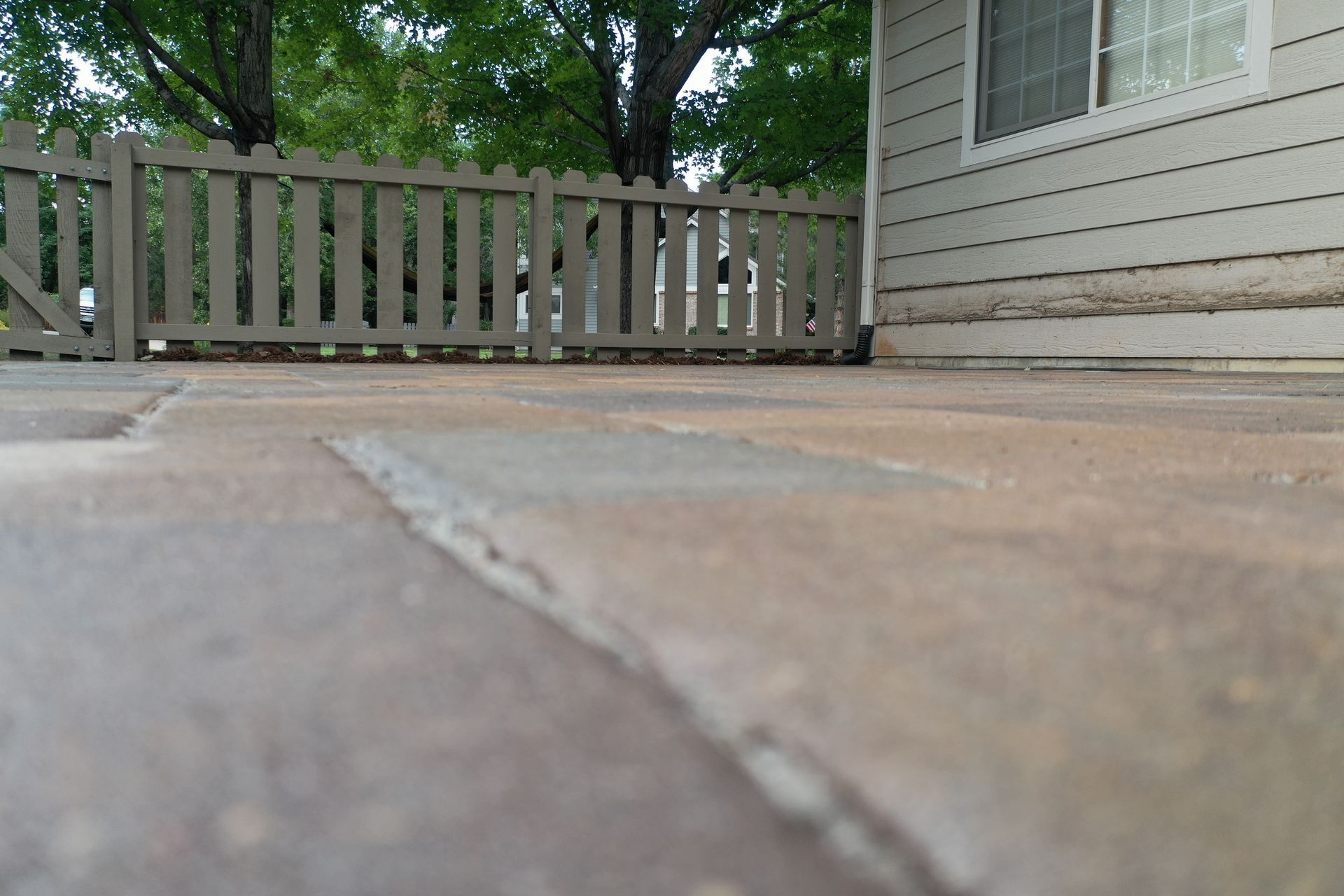 Close-up view of brick pavers on a patio, with a wooden fence and siding of a building visible in the background.