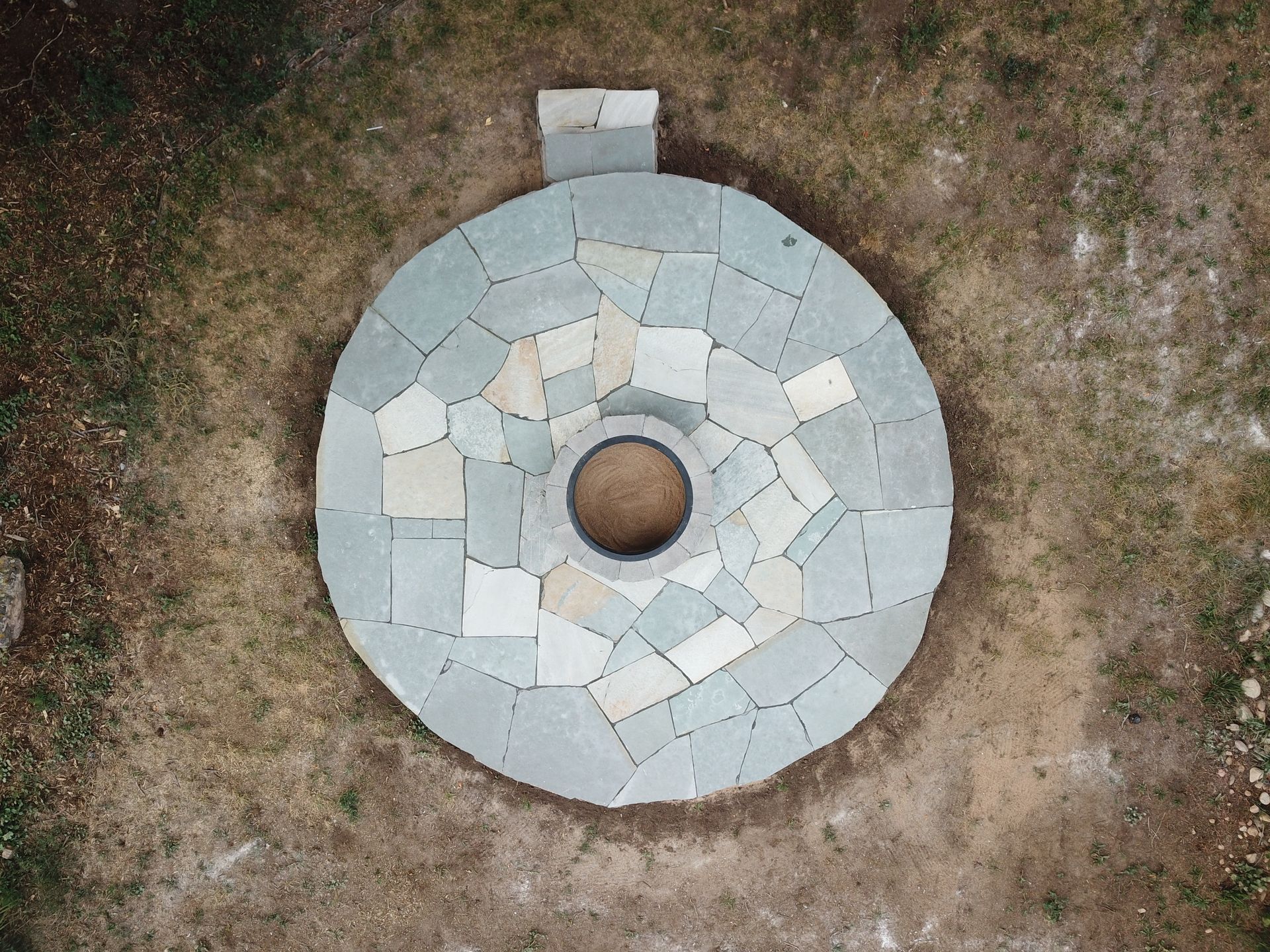 Overhead view of a stone-paved fire pit in a grassy area with a small chimney feature.