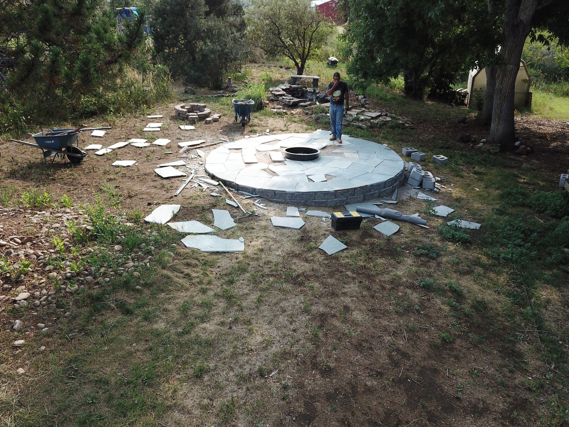 Man building a stone patio in a grassy yard, surrounded by tools and materials.