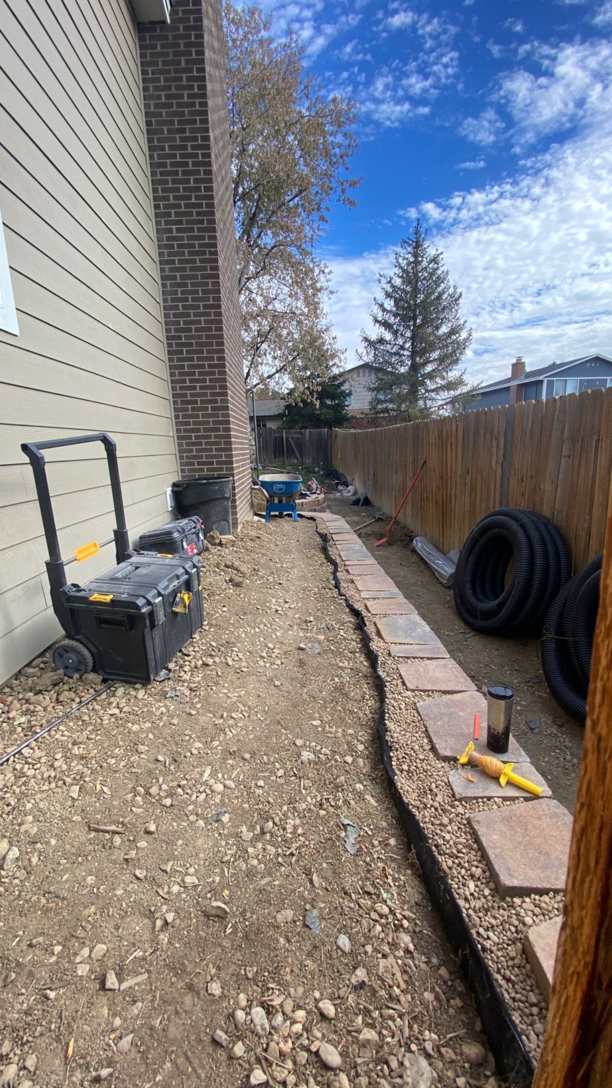 Side yard with brick wall, path, fence, and tools. Dirt ground with a blue sky.