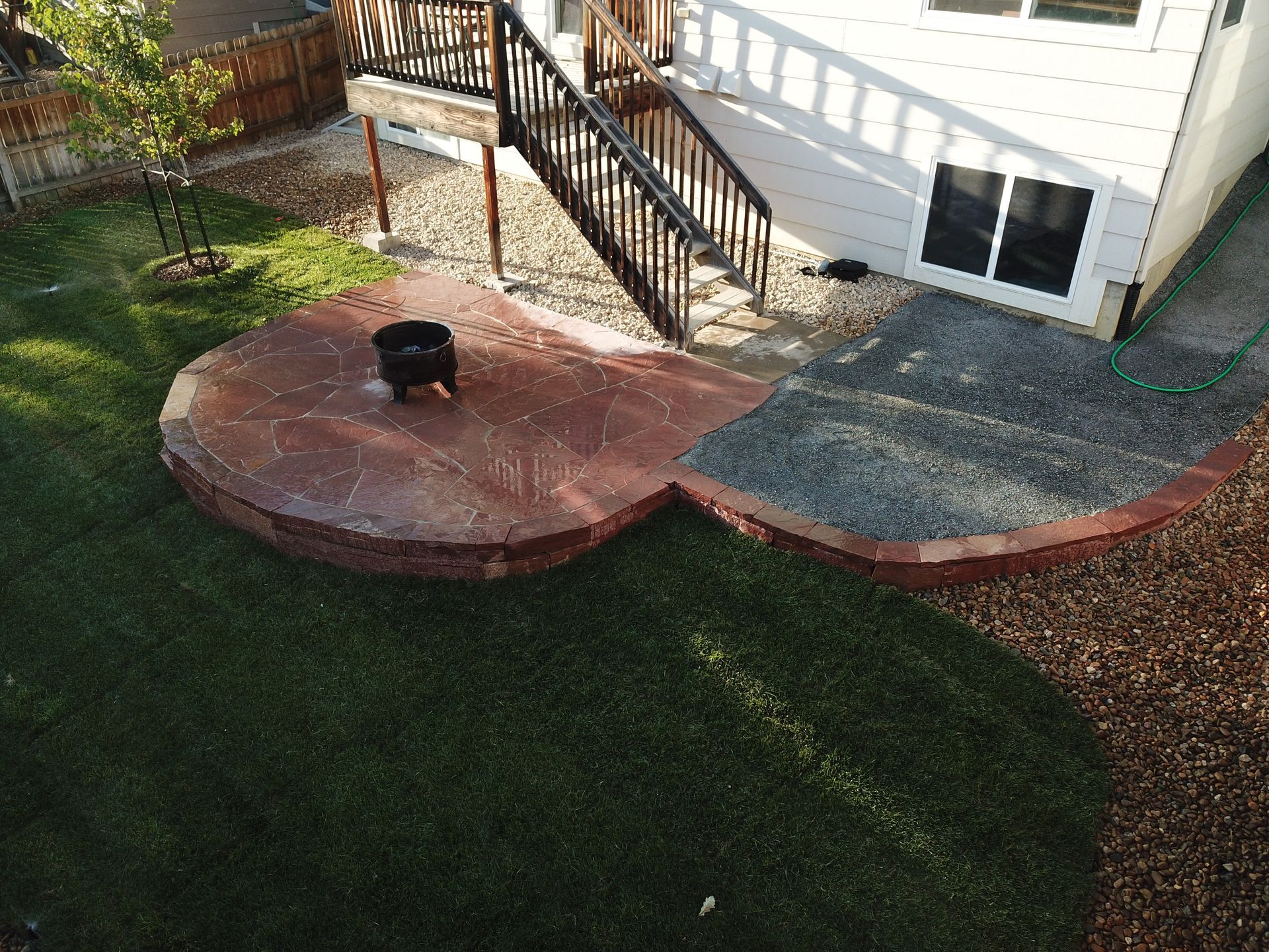 Backyard patio with fire pit, bordering lawn and gravel against a white house with stairs.