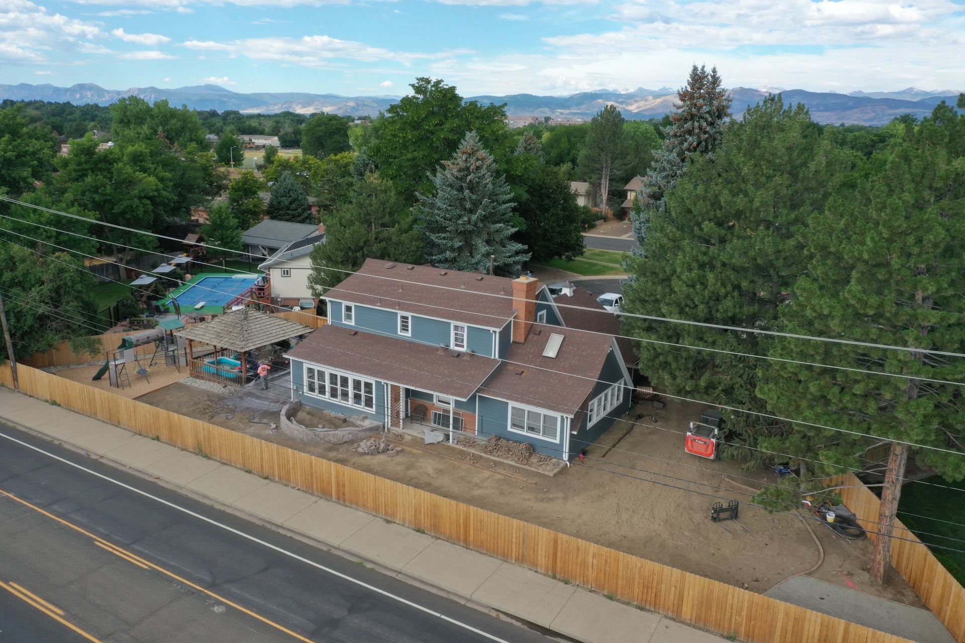 Aerial view of a blue two-story house with a wooden fence, trees, and mountains in the background.