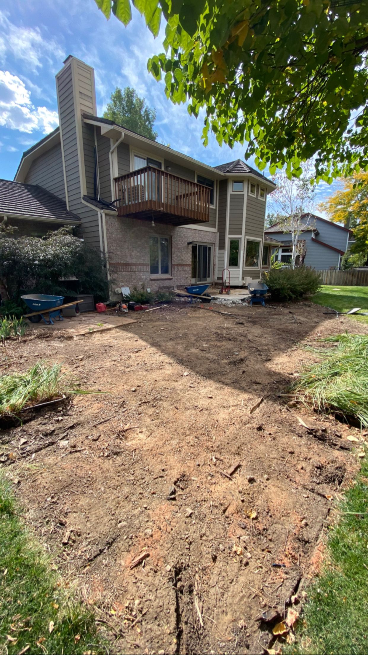 Backyard with dirt patch, house with deck, and chimney under a blue sky.