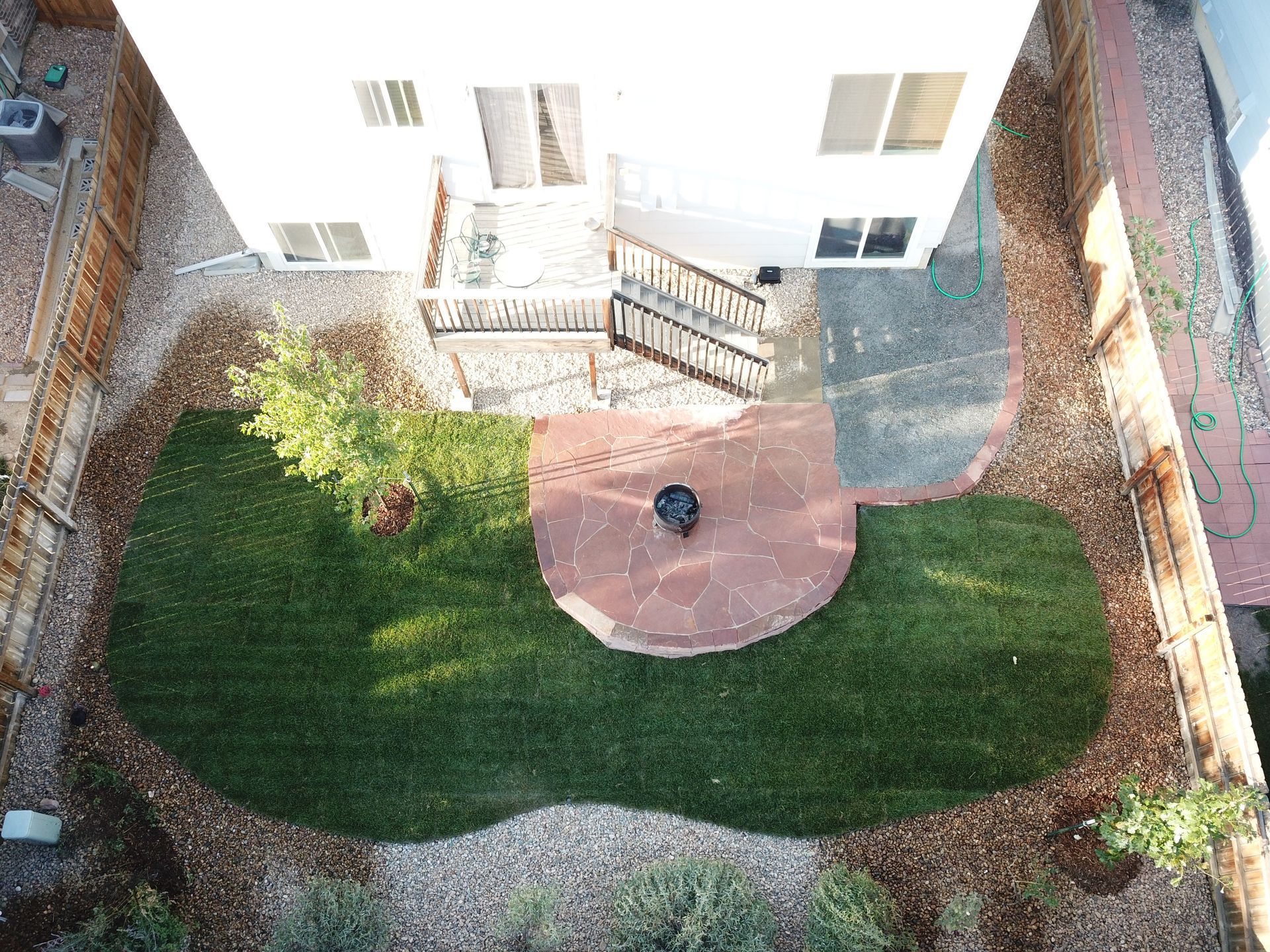 Overhead view of a backyard with green grass, a red stone patio with a fire pit, and a wooden deck connected to a white house.