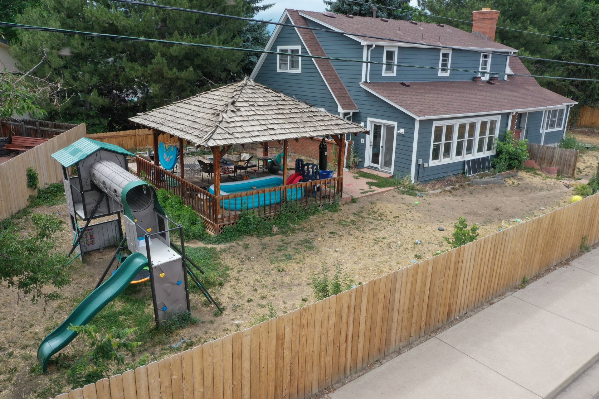 Backyard with blue house, brown gazebo over a pool, wooden fence, and a playground.