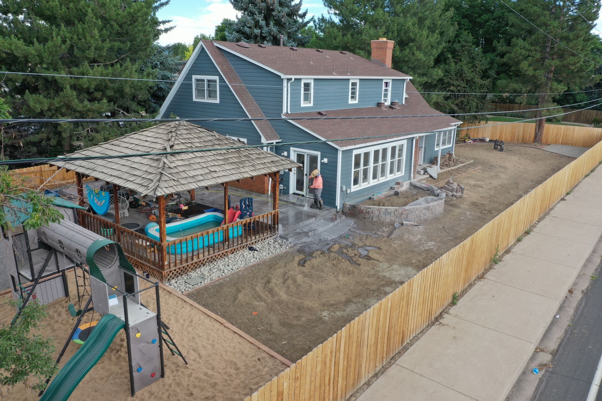 Backyard with blue house, brown gazebo with hot tub, playground, and wooden fence.