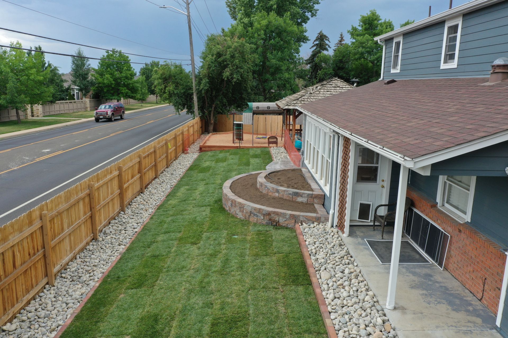 Aerial view of a home with a wooden fence, green lawn, a brick patio, and a road.