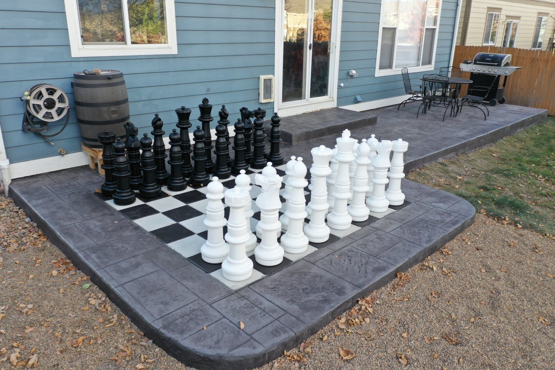 Giant chess set on a concrete patio next to a house with a barrel and grill. Black and white pieces.