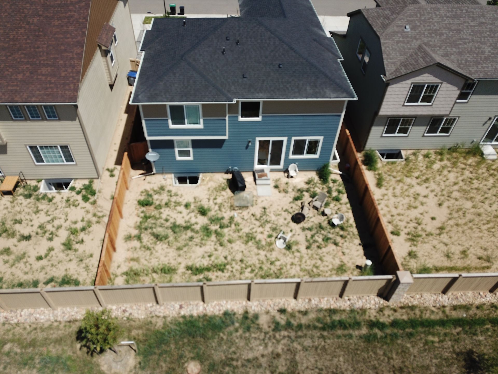 Backyard view of a two-story blue house with sparse grass, brown fences, and neighboring houses.