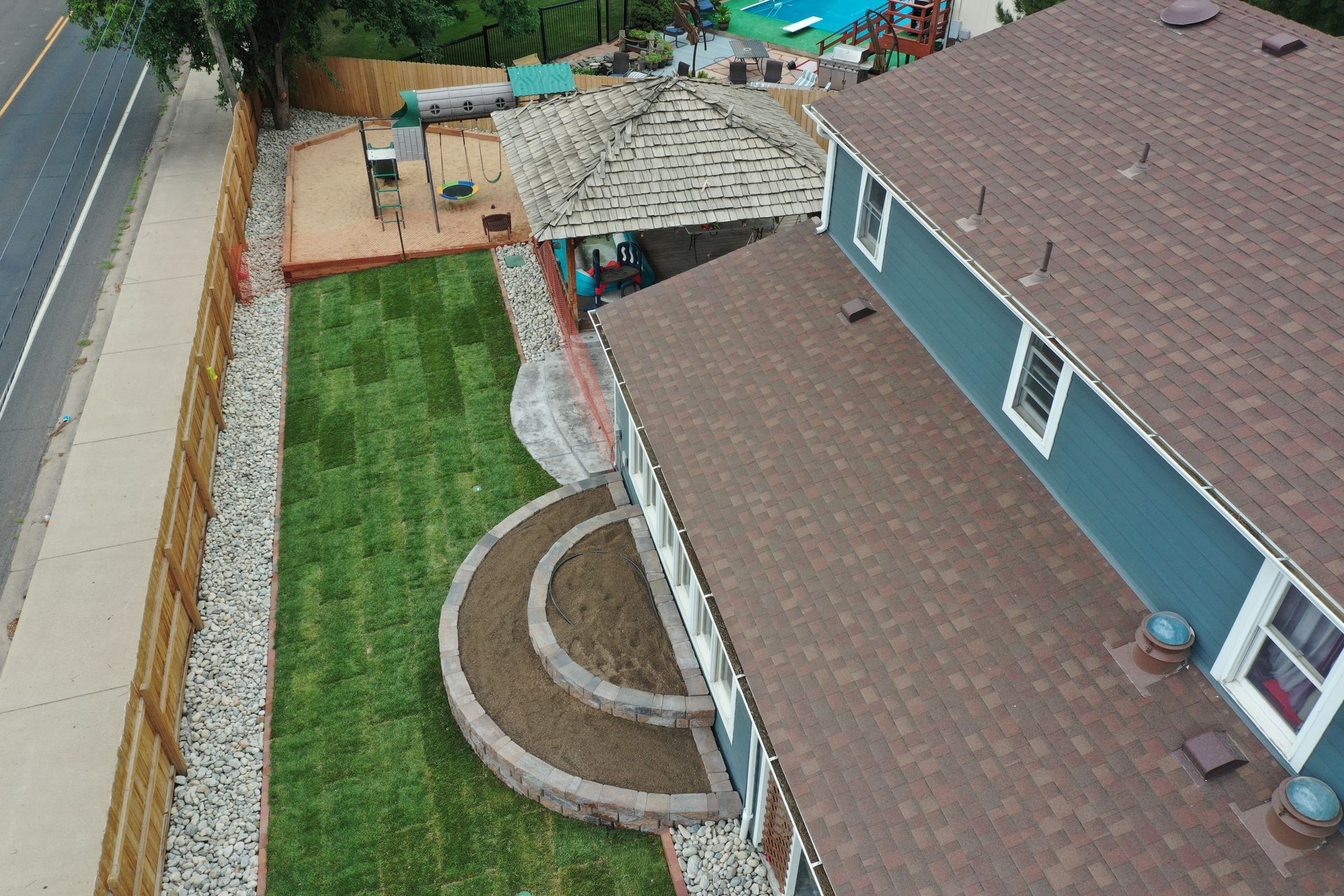 Aerial view: Blue house with brown roof and green lawn, next to a road and a wooden fence.