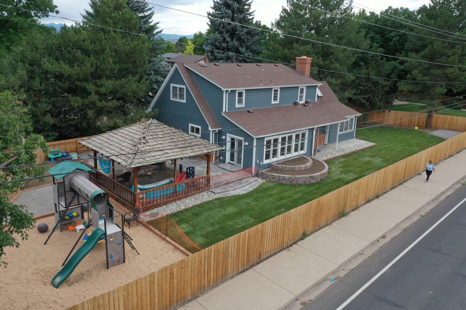 Two-story blue house with a play set, surrounded by a wooden fence, next to a road, and a person walking by.