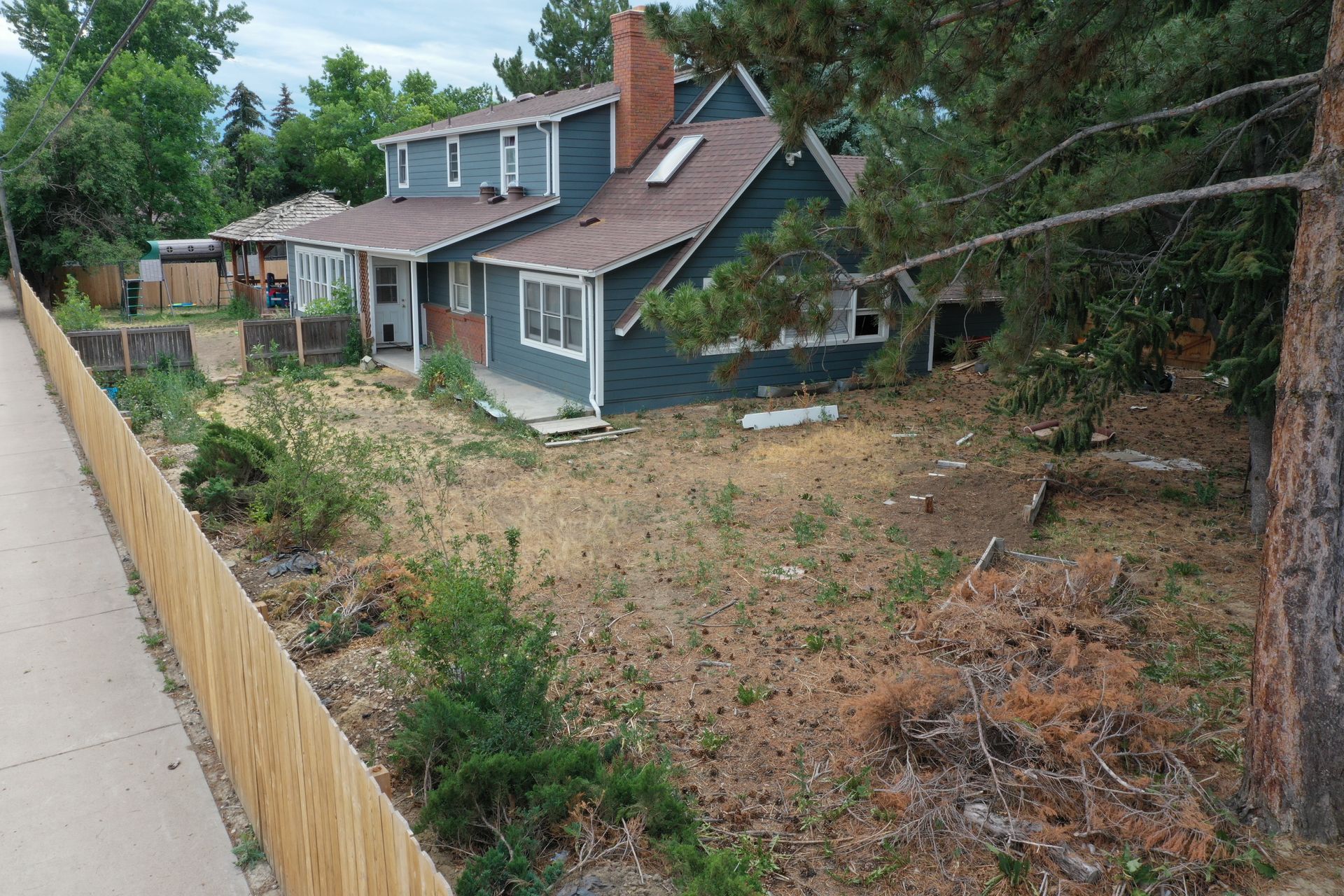 Two-story blue house with dry yard, wooden fence, and tall trees.