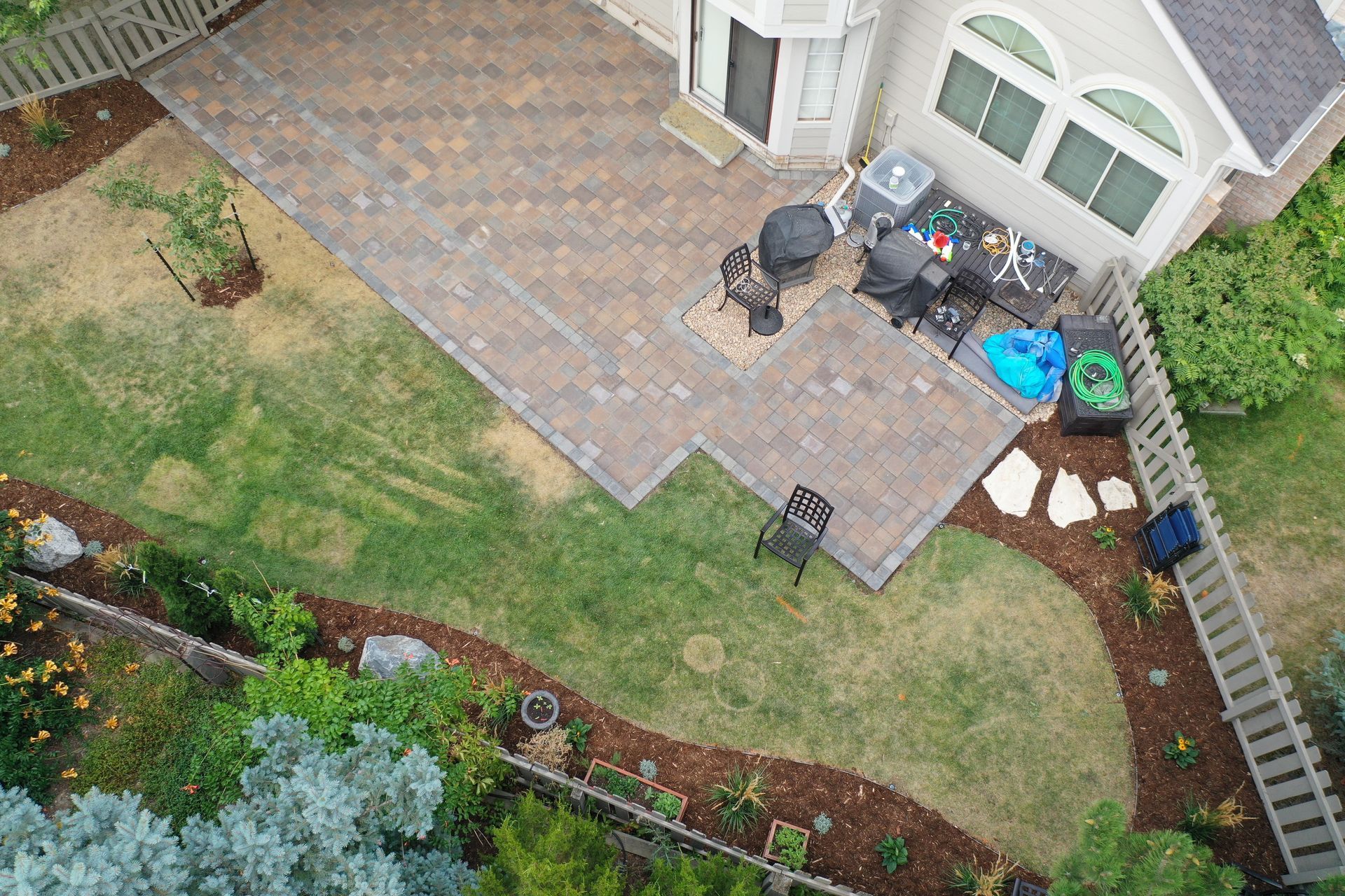Overhead view of a brick patio with grill, table, and chairs near a house and grassy yard.