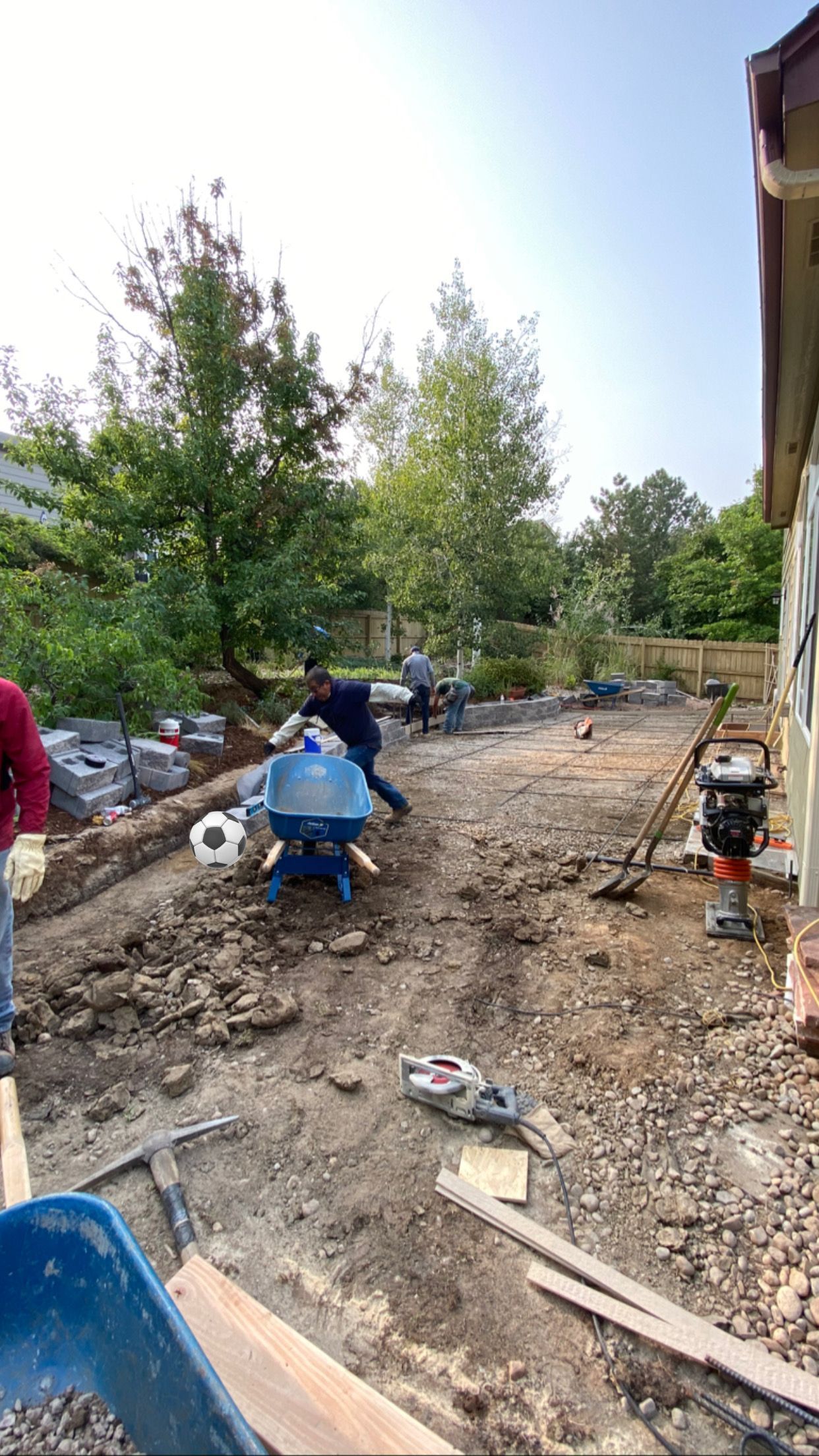 Construction workers laboring in a dirt yard near a building, using tools and equipment, on a sunny day.
