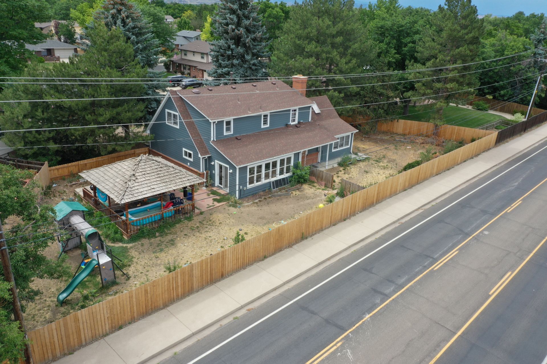 Two-story blue house with brown roof and wooden fence beside a road.